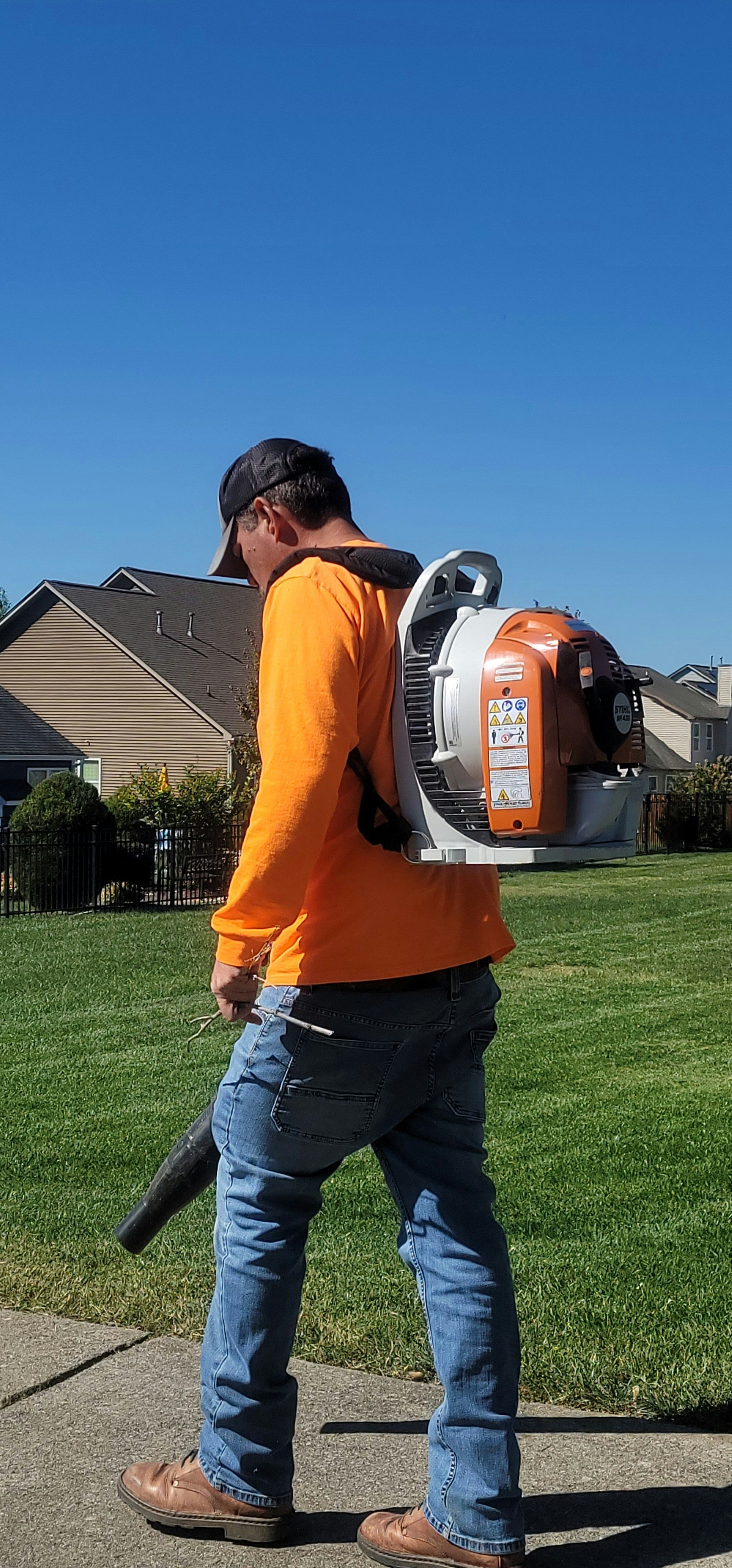 Man in orange shirt wearing a backpack leaf blower while walking on a sidewalk in a residential neighborhood.