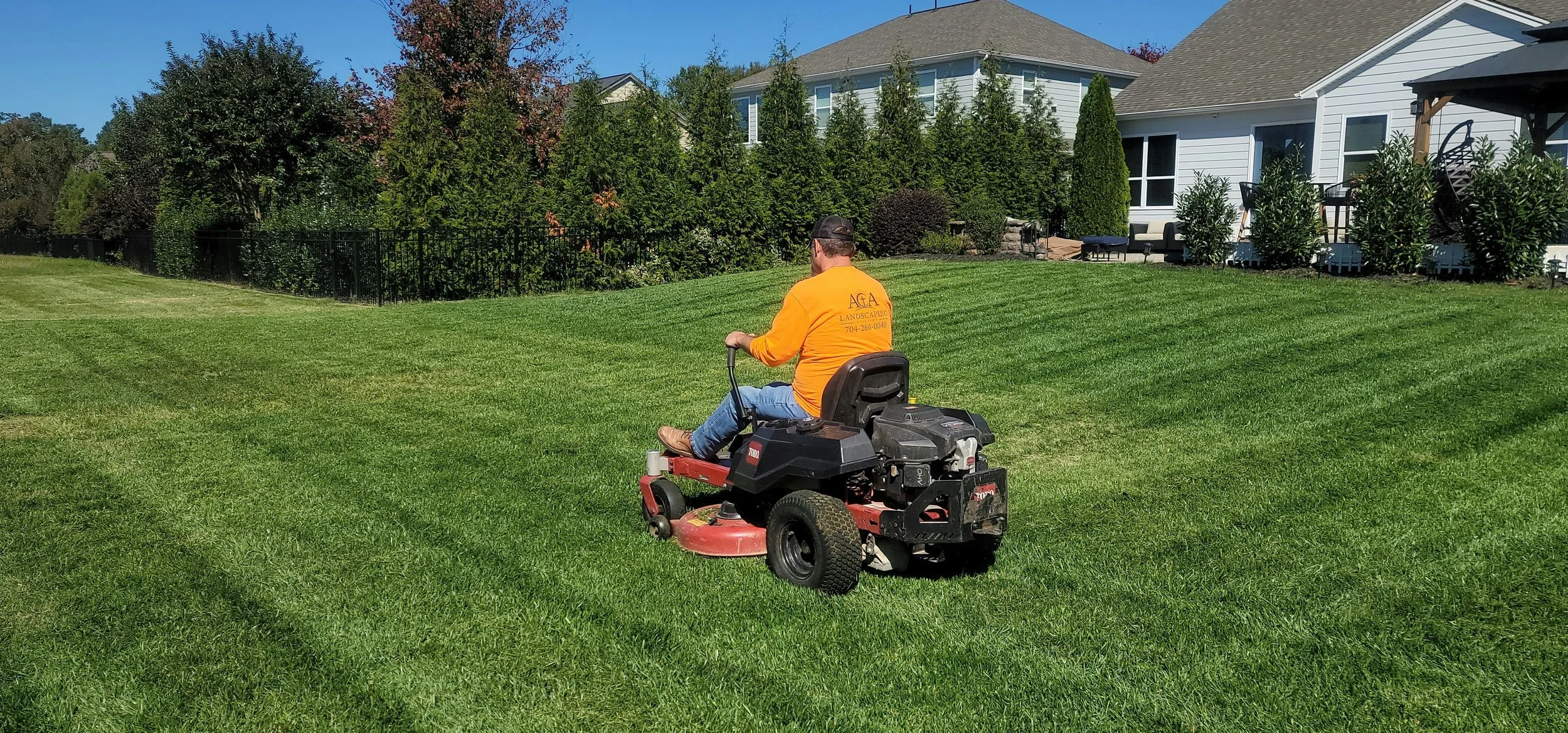 A person wearing an orange shirt and blue jeans mowing a well-maintained green lawn with a riding lawn mower. In the background, there are houses, trees, and shrubs, under a bright blue sky.
