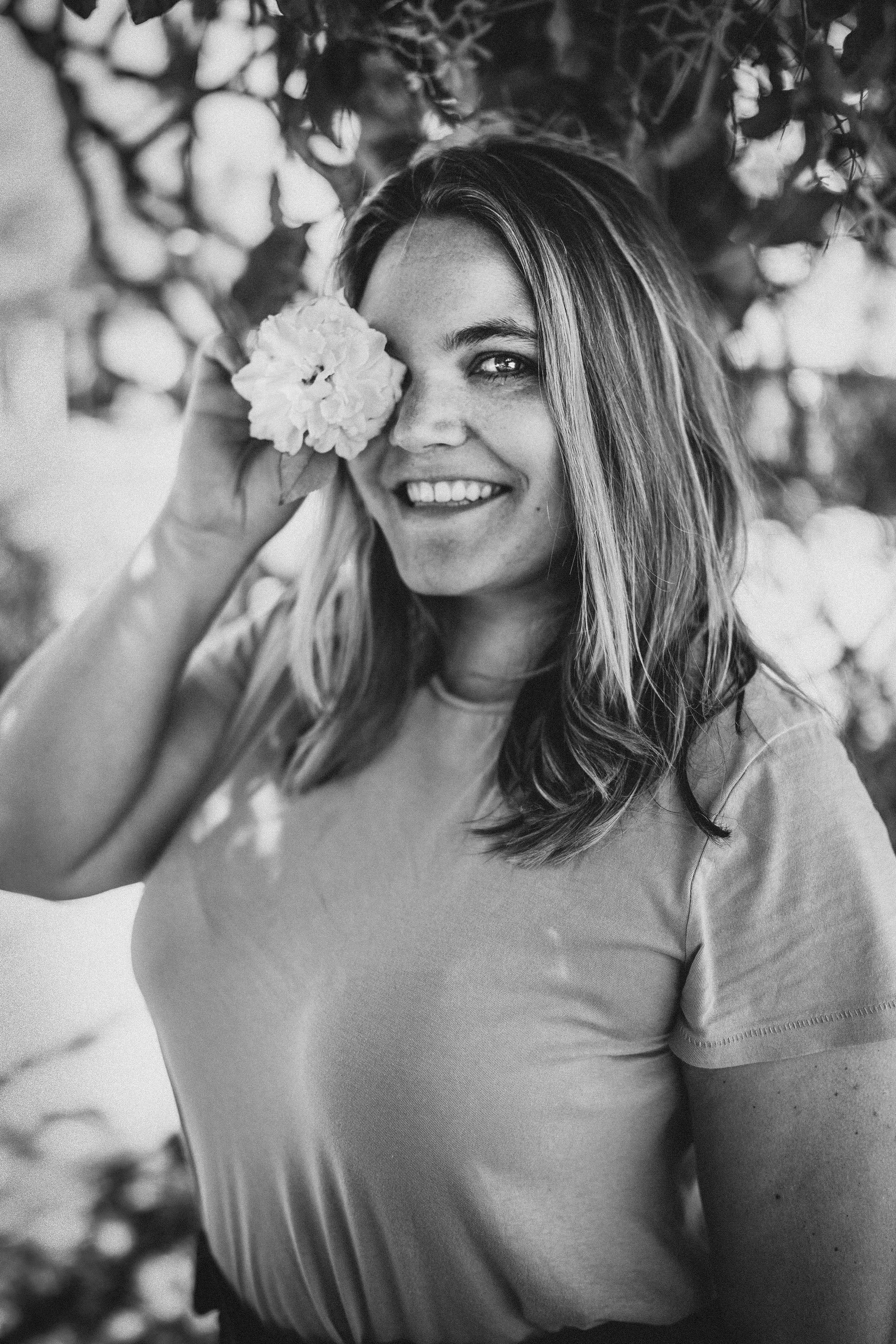 A smiling woman with long hair holding a flower over her eye, standing outdoors with blurred flowers and foliage in the background.