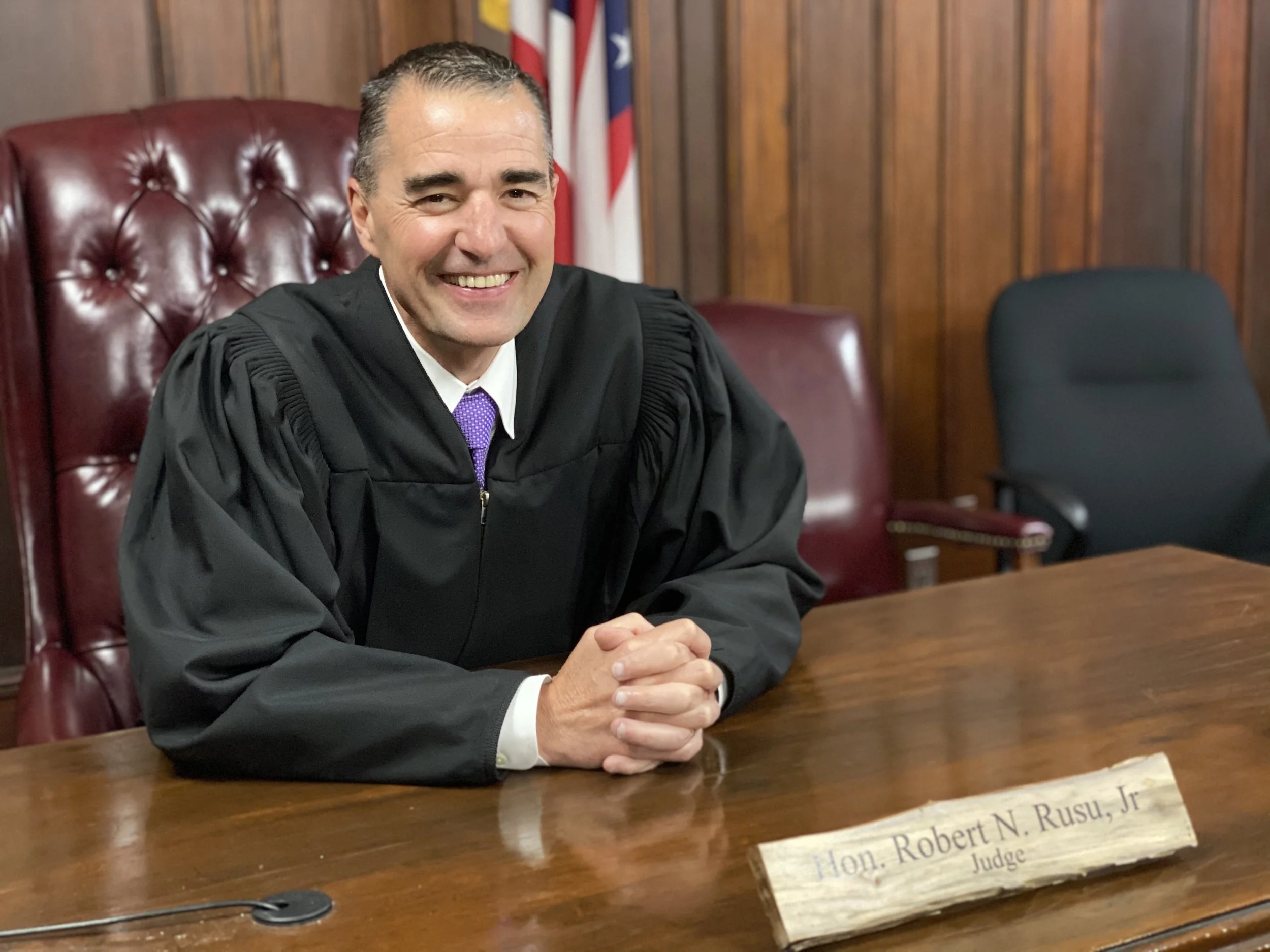 A judge wearing a black robe sitting at a wooden judge's bench, smiling, with a wooden nameplate that reads "Hon. Robert N. Rusu, Jr. Judge" in front of him, in a courtroom with wood-paneled walls and American flags in the background.