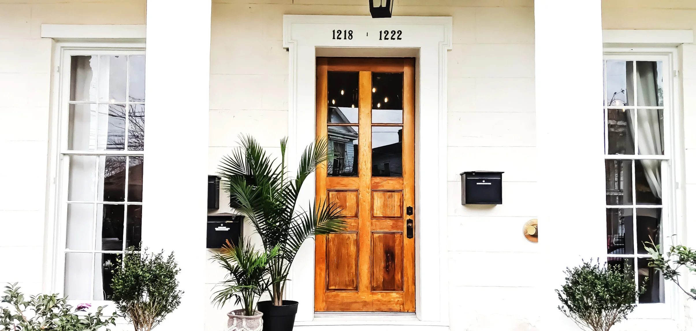 Front door of a house with a wooden door, two windows on each side, and house numbers 1218 and 1222 above the door. There are plants in pots and black mailboxes on either side of the door.