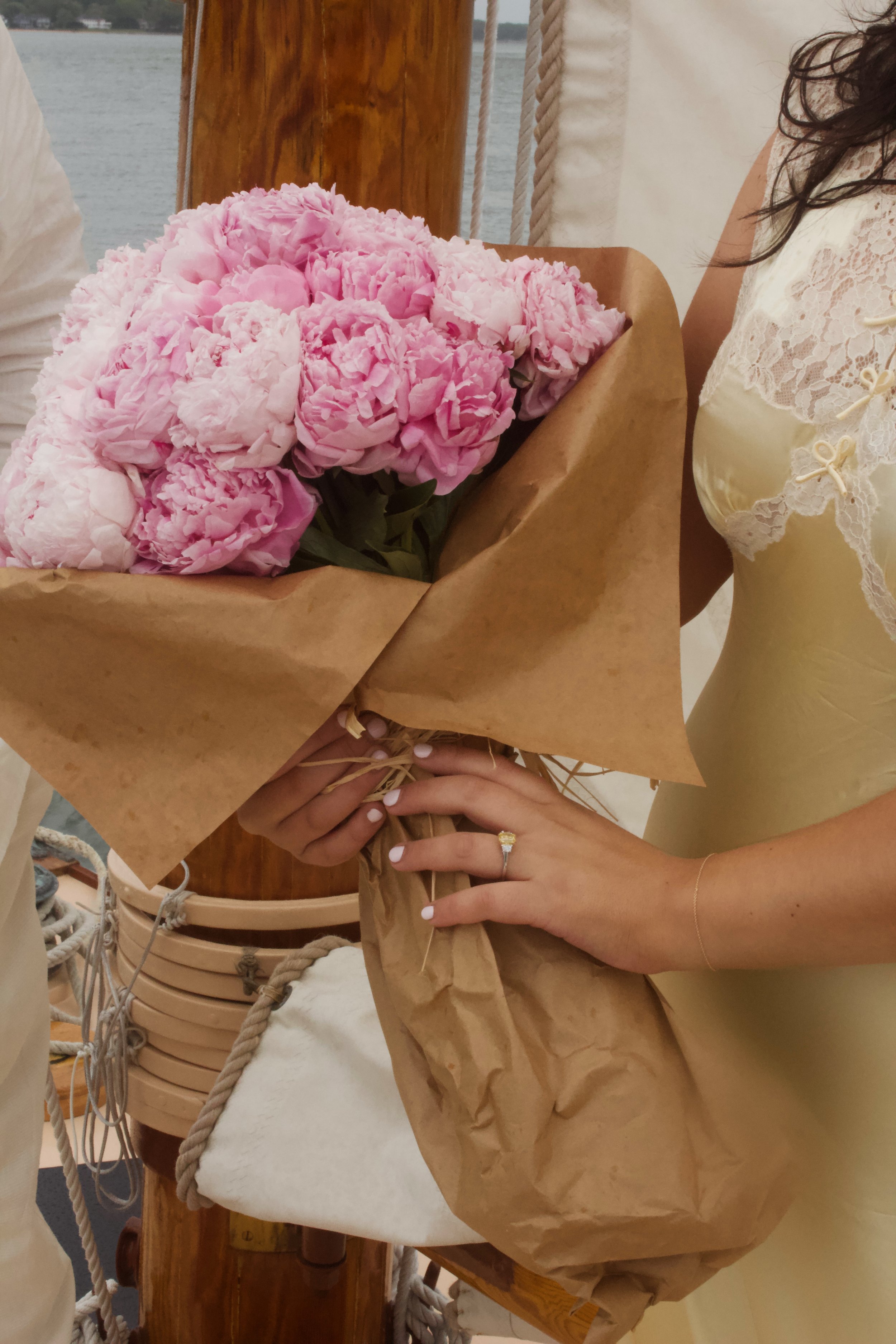 Person in yellow dress holding a large bouquet of pink peonies wrapped in brown paper on a wooden table near water.