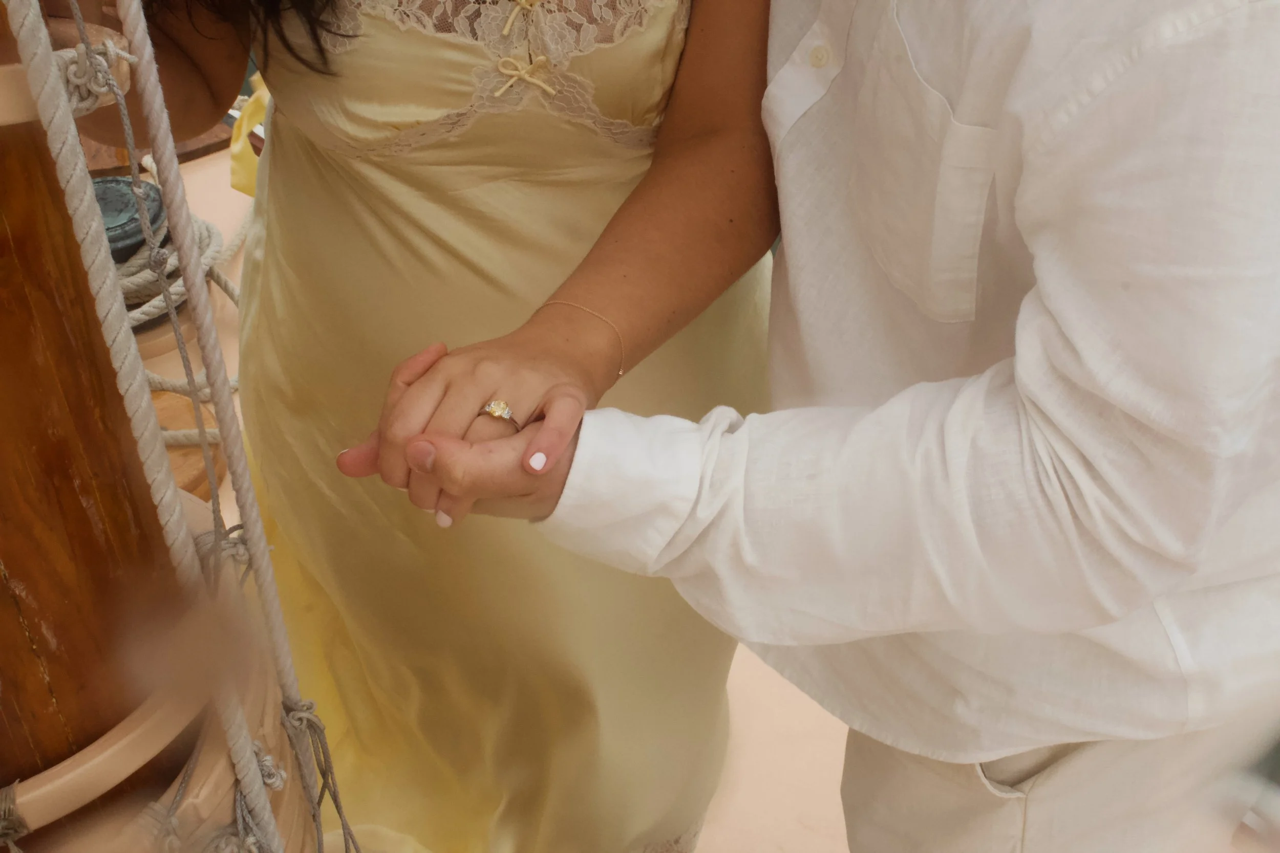 An elderly couple holding hands, with the woman wearing a yellow lace nightgown and a wedding ring, sitting together in bed.