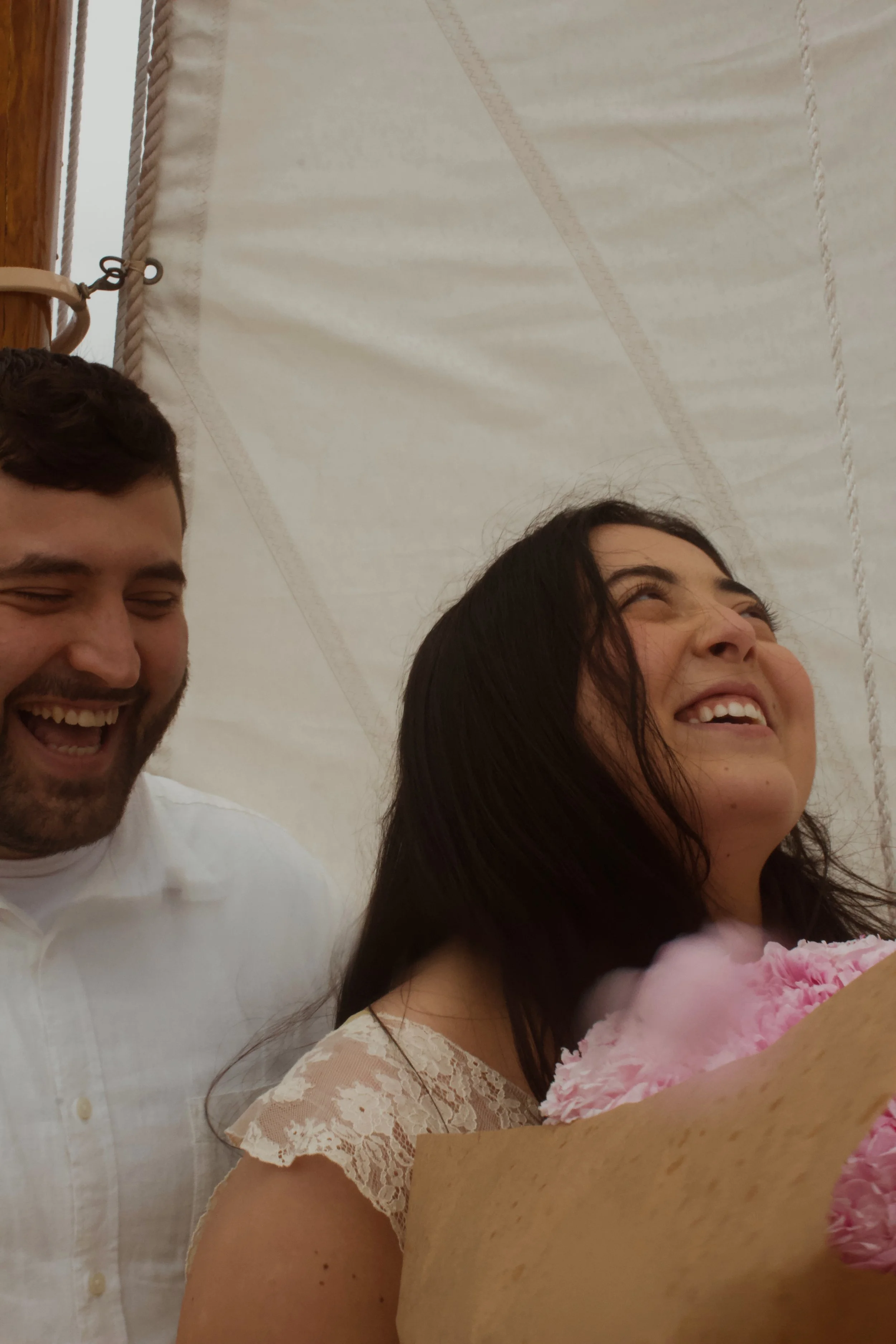 Two women and a man are smiling and laughing, standing in front of a white tent, with one woman holding a bouquet of pink flowers.