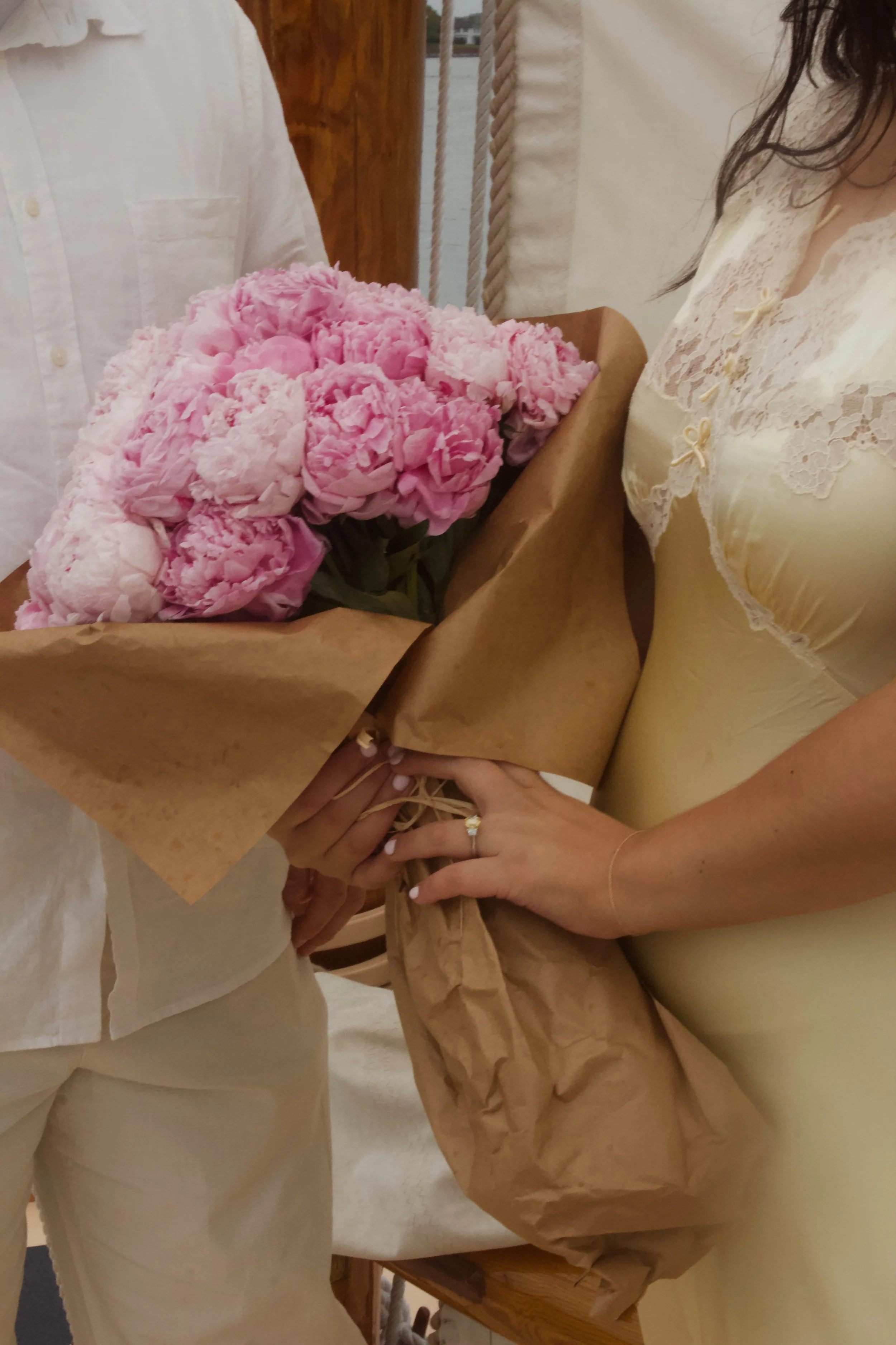 Two women exchanging a large bouquet of pink peonies, one woman is wearing a cream lace dress, and the other is dressed in white.