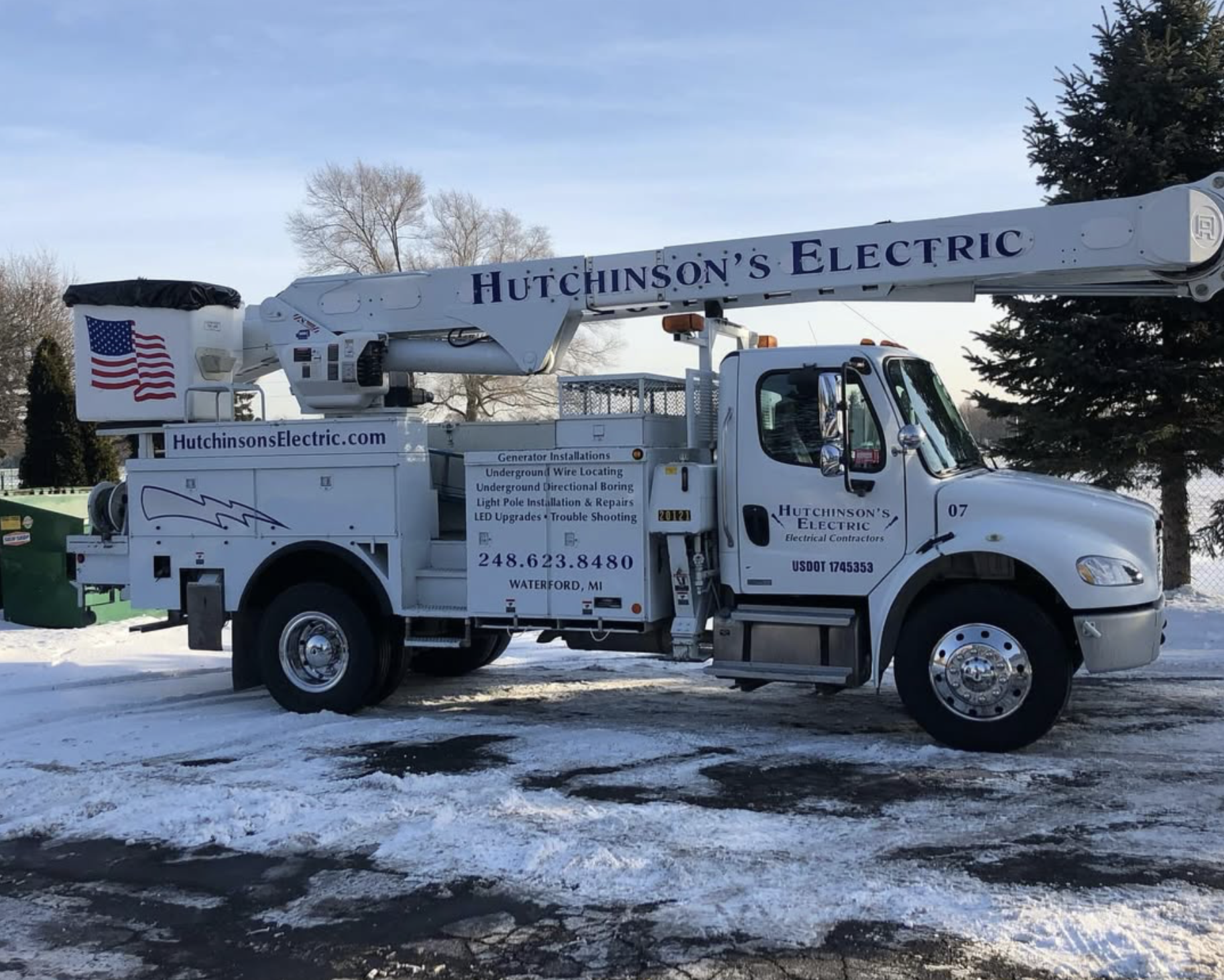 A white Hutchinson's Electric utility truck with a crane and an American flag on the side parked on snow. The truck has company contact information and services listed on it, including generator installations and underground wire locating.