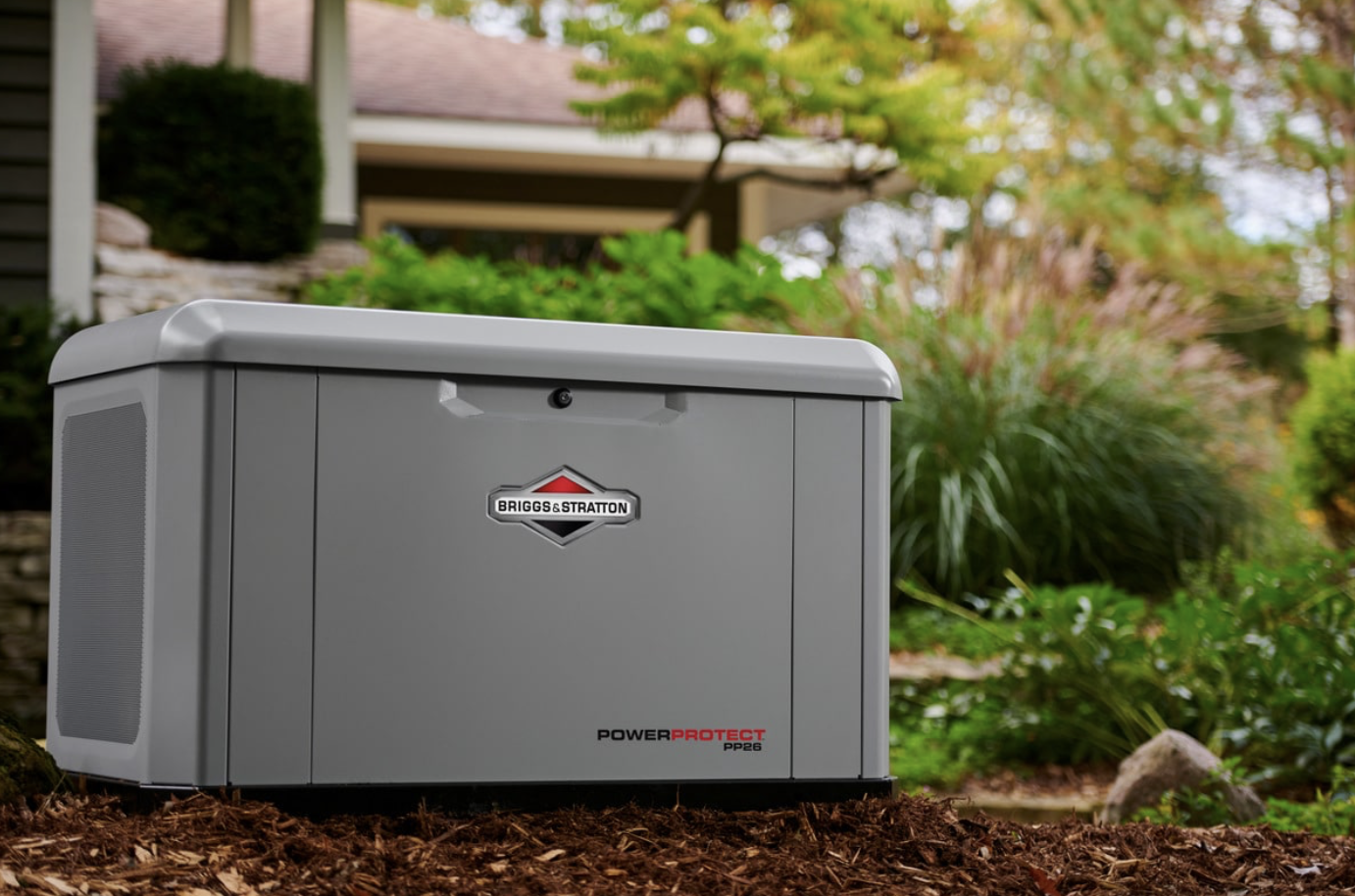 A grey Briggs & Stratton PowerProtect PP26 outdoor generator or compressor outside in a garden area with green bushes and a house in the background.