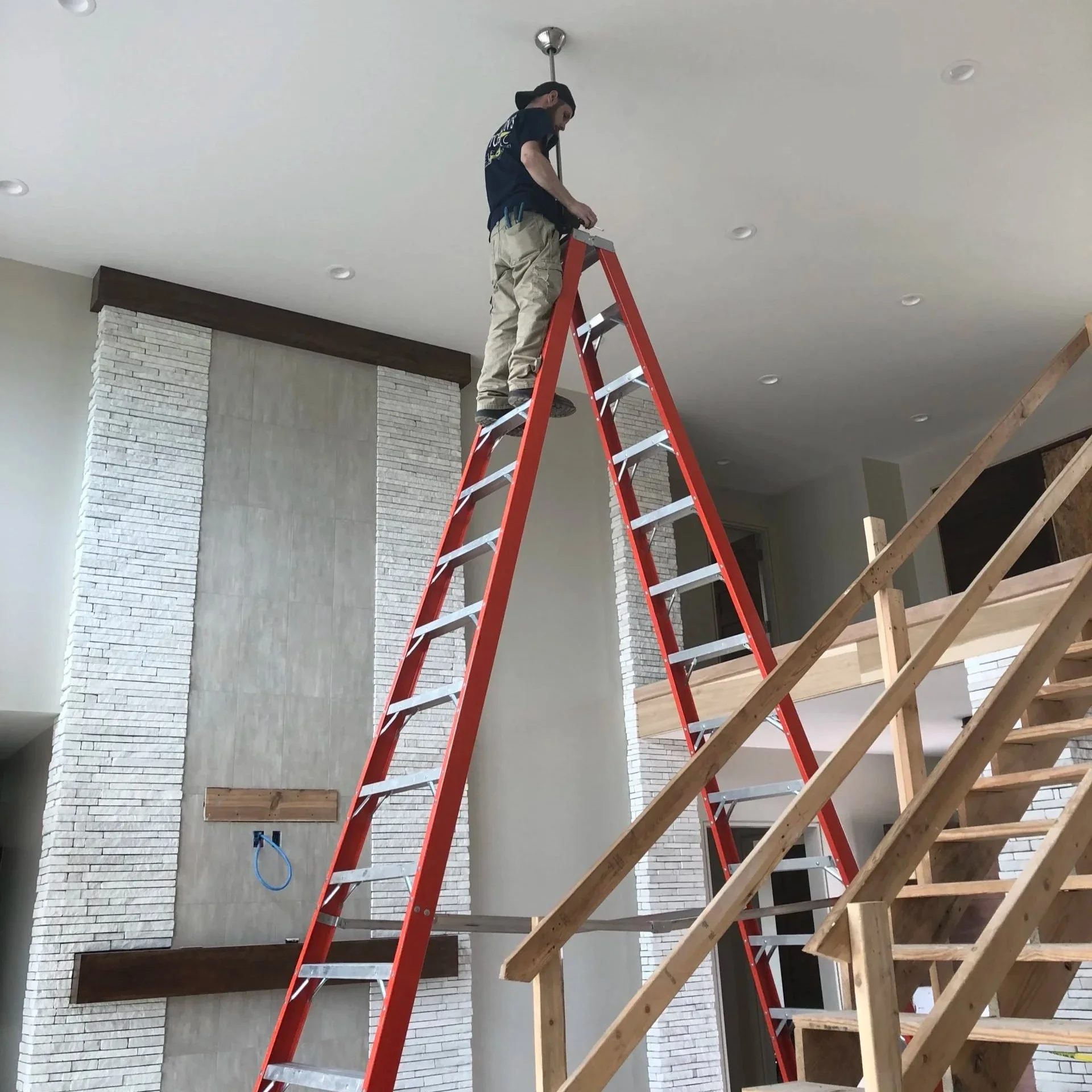A man standing on top of a tall red ladder inside a house, working on the ceiling. The house has a modern design with white brick and concrete walls, and an unfinished wooden staircase.
