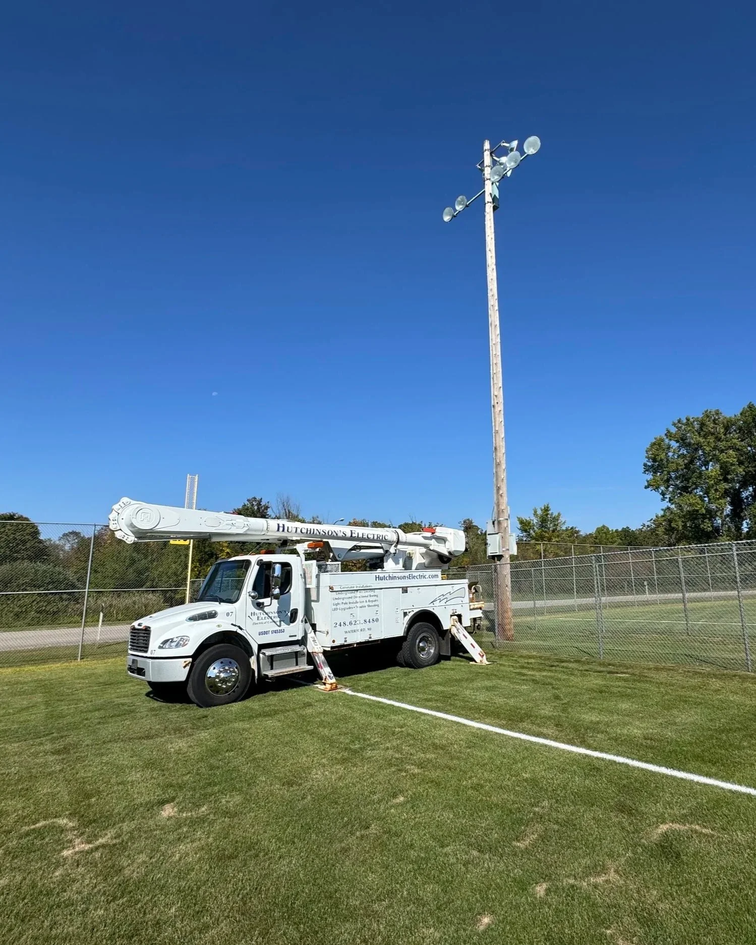 A utility truck from Hutchinson's Electric parked on a grassy field near a tennis court, with a tall wooden utility pole and a clear blue sky in the background.