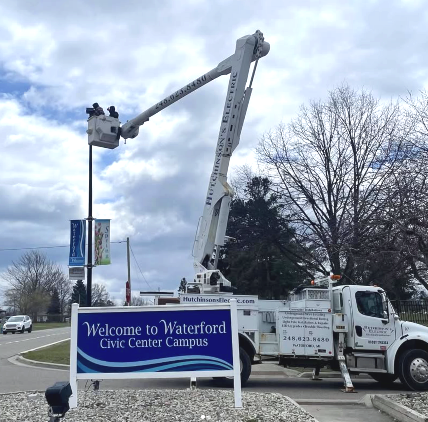 Elevated workers installing or maintaining a streetlight at Waterford Civic Center Campus, with a large truck and an inspection bucket lift visible, and a sign welcoming visitors.