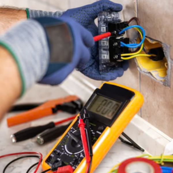A person wearing blue gloves using a multimeter to test electrical wires connected to a circuit breaker inside an electrical socket on a wall with tile. The multimeter displays a value of 0.00.