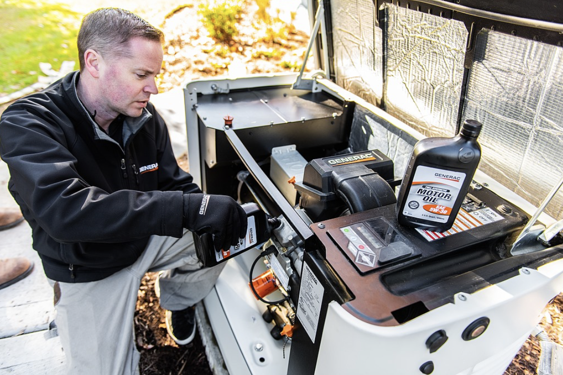 Man checking or servicing a generator outdoors with a bottle of motor oil nearby.