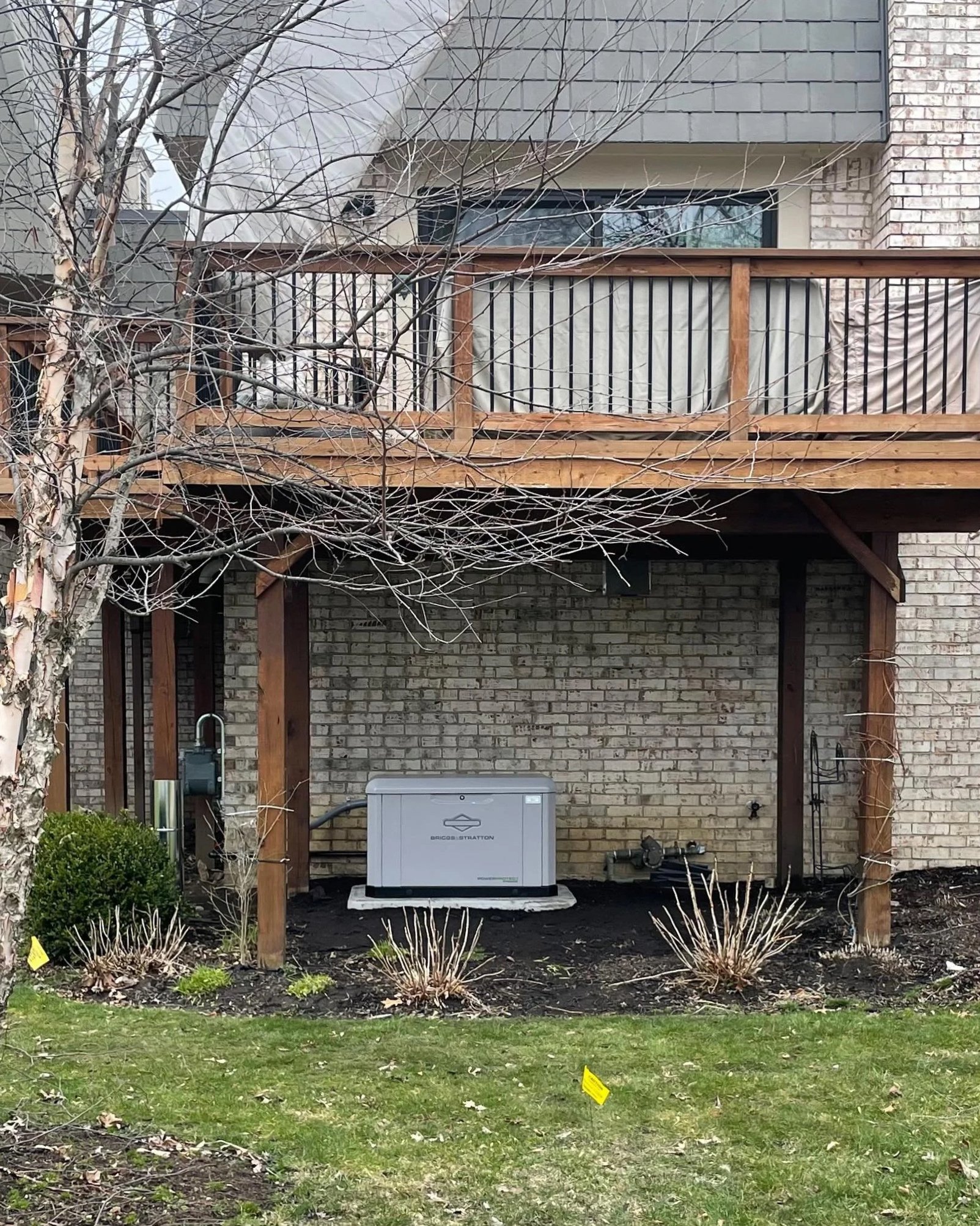 Backyard with a wooden deck, a brick wall, leafless tree, and a green lawn with small plants. A grey condensation-style air conditioning or heat pump unit is placed underneath the deck, with some utility hookups nearby.