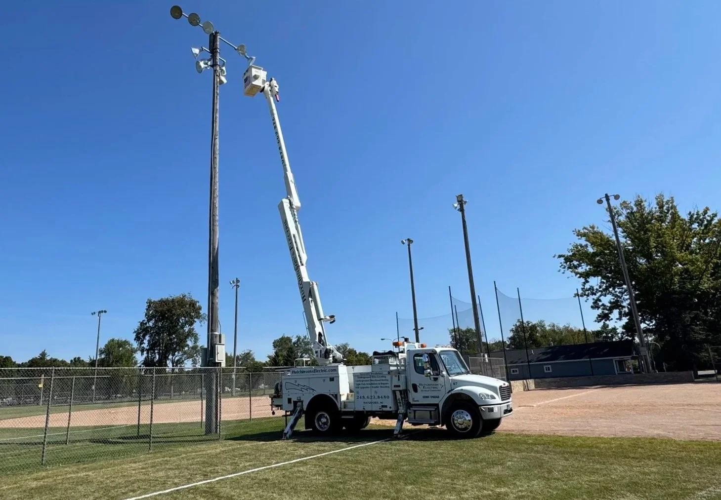A utility truck with an extendable crane reaching a tall utility pole at a sports field with a chain-link fence, trees, and baseball fields.