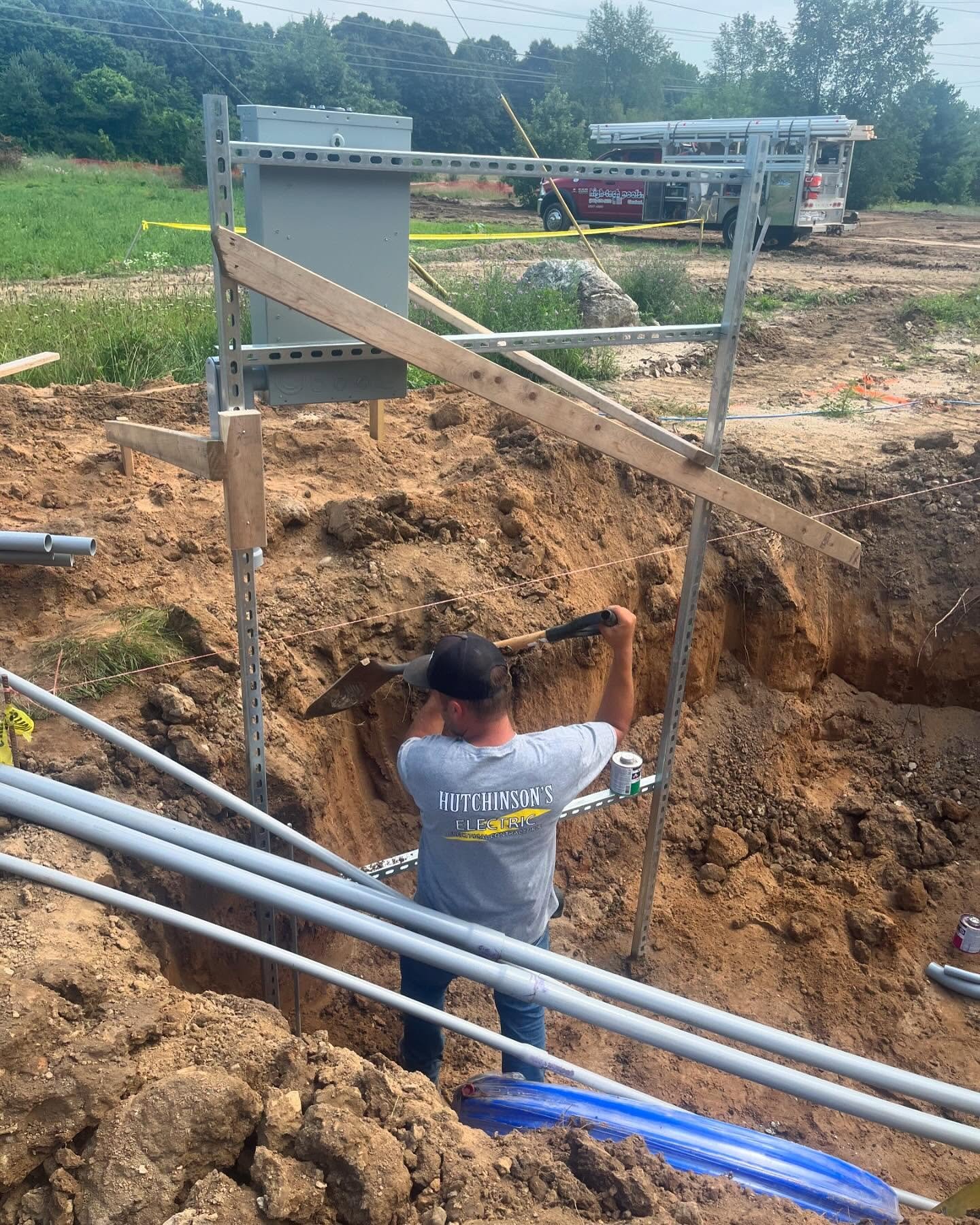 A construction worker digging a trench with a shovel at an outdoor construction site. The worker is wearing a gray T-shirt with the words "HUTCHINSON'S ELECTRIC" on the back, and a black cap. There are steel and wooden support structures and pipes around the excavation site, with a utility truck and trees in the background.