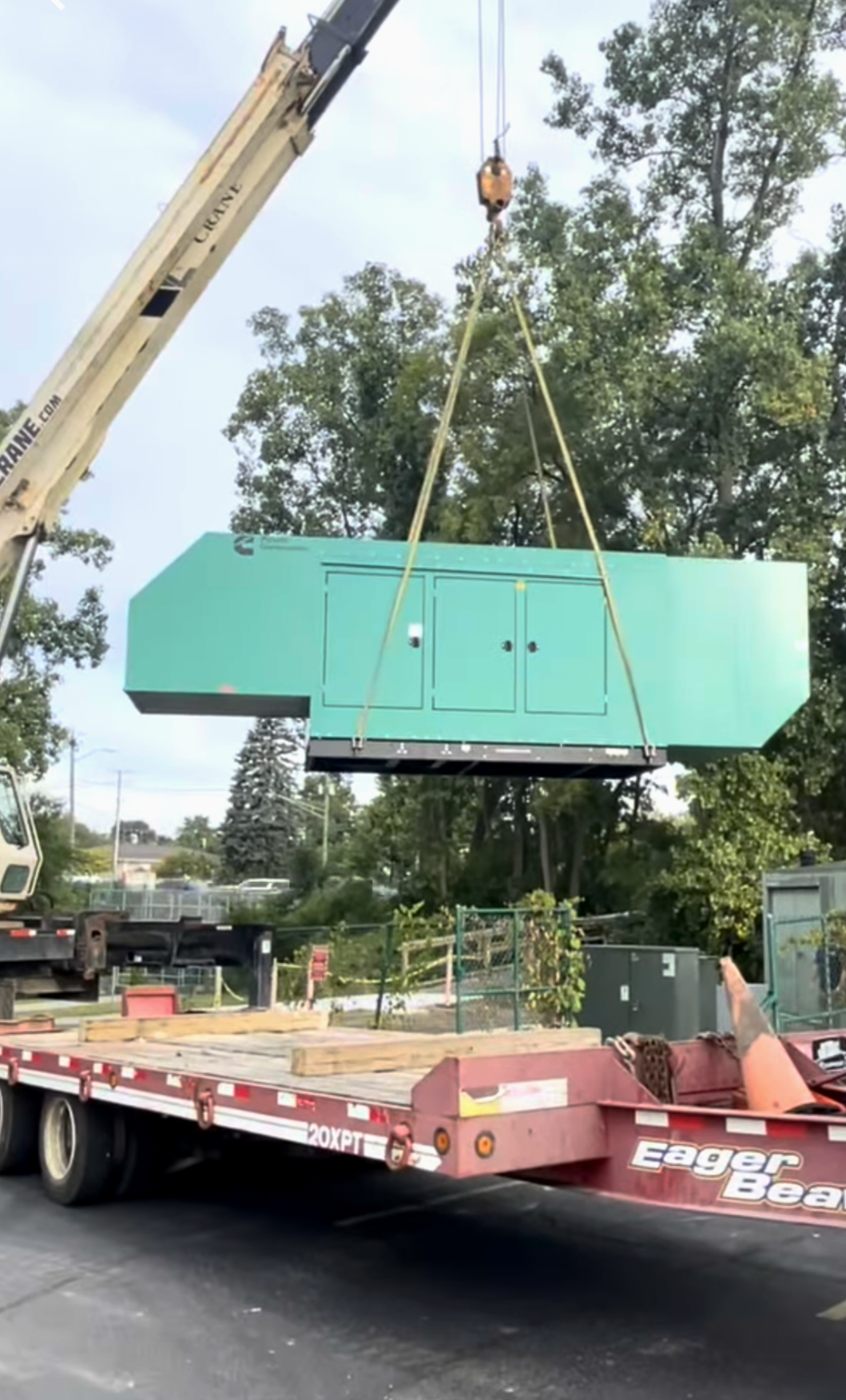 A construction crane lifting a large green industrial unit onto a flat trailer in a parking lot surrounded by trees.