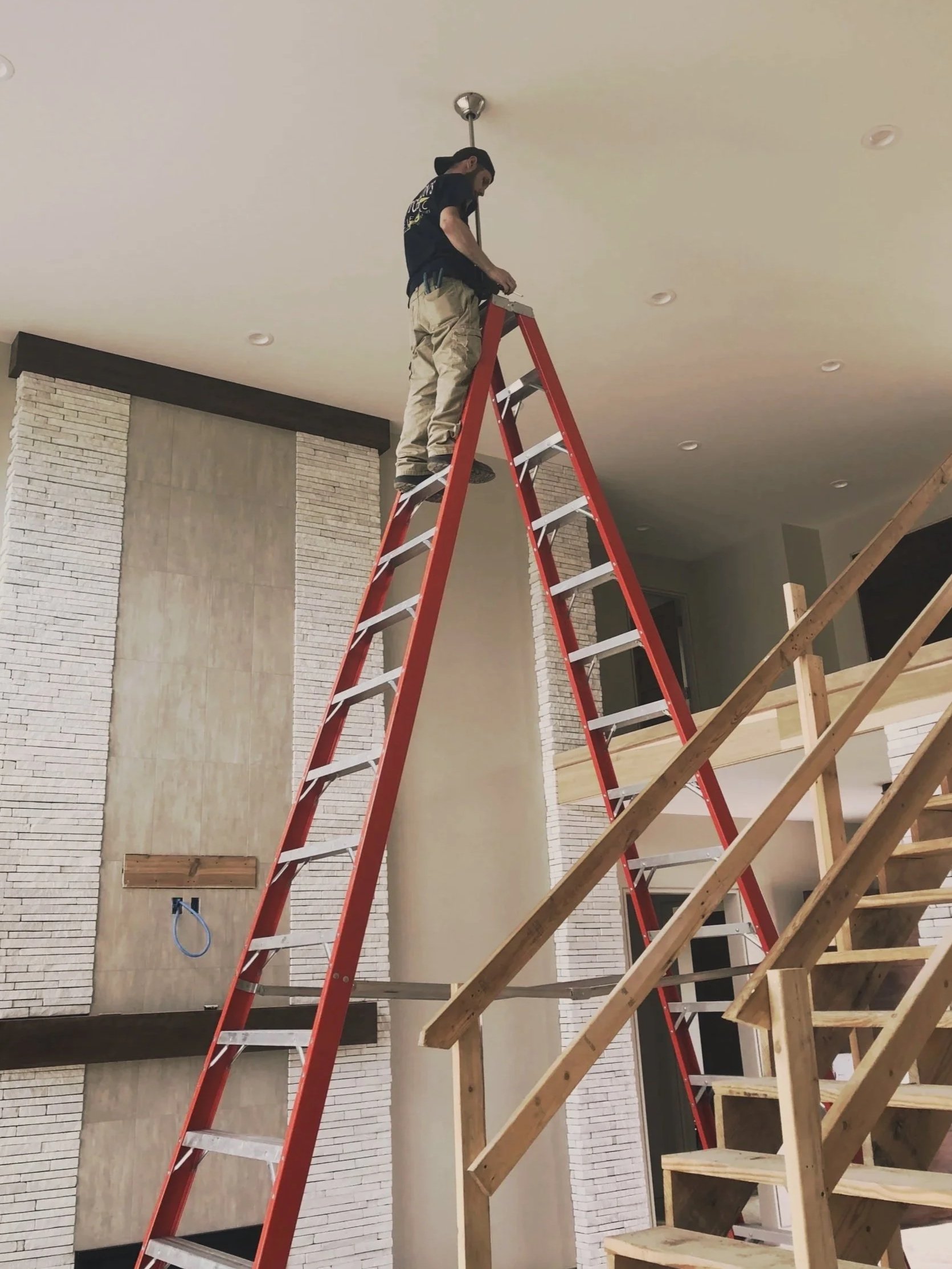 Electrician standing on a tall ladder working on a ceiling lighting installation.
