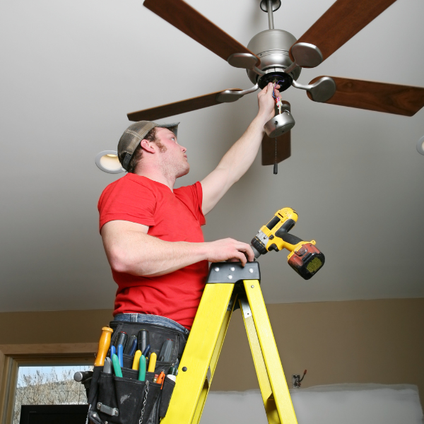 A man wearing a red t-shirt and a baseball cap standing on a yellow ladder, installing or fixing a ceiling fan while holding a cordless drill.