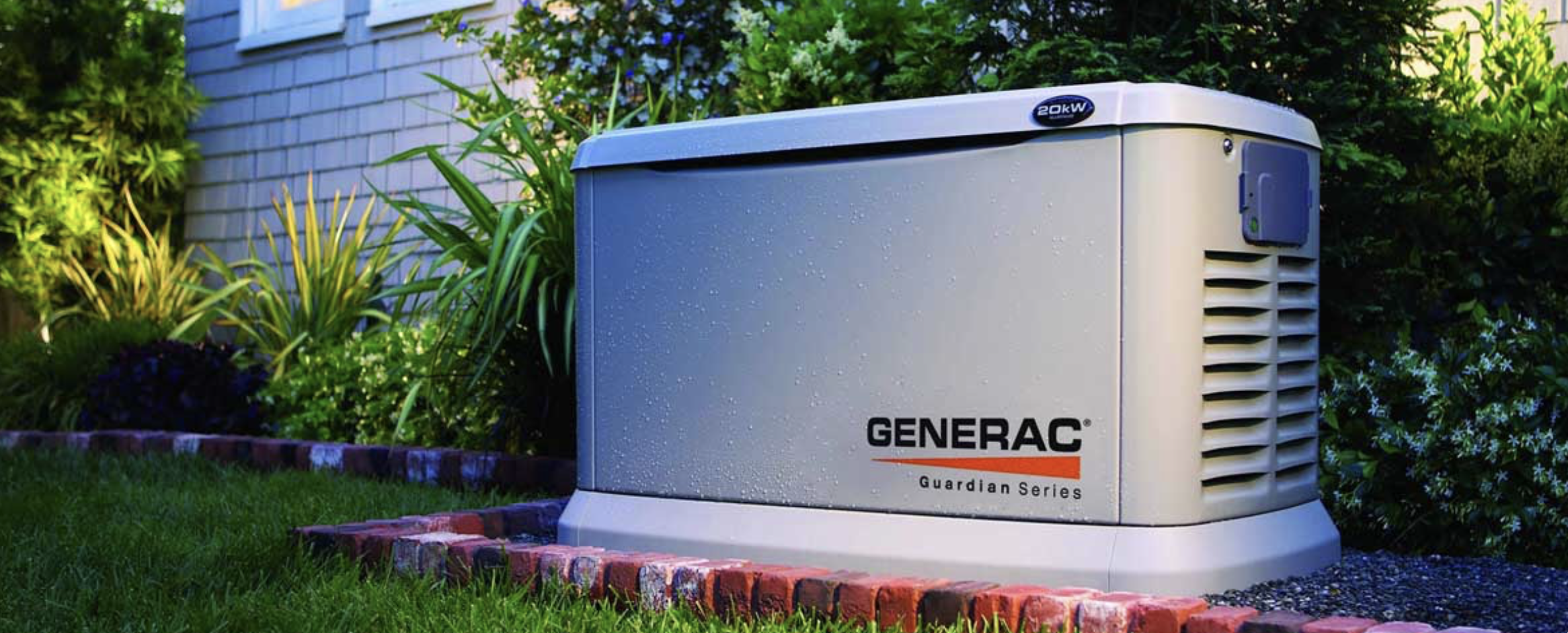 A Generac Guardian Series home standby generator installed outdoors on a concrete pad next to a garden bed with plants and shrubs.