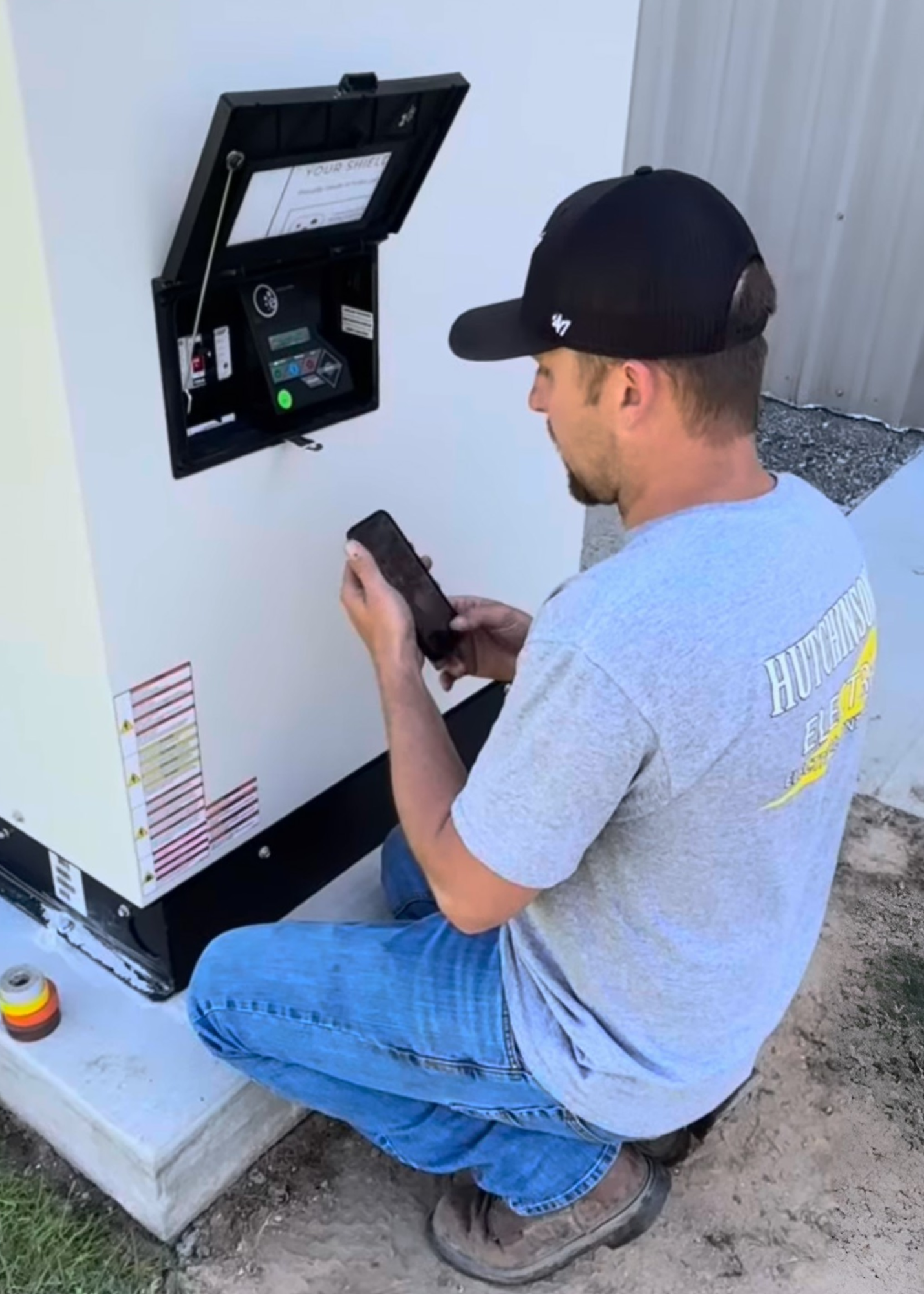 A young man wearing a black cap, gray t-shirt, and blue jeans crouching outside and using his smartphone near a white utility box. The box has an open panel with electronic components inside and a menu displayed on the ceiling. There is a small round tool or sensor with yellow, red, and black stripes resting on the ground nearby.