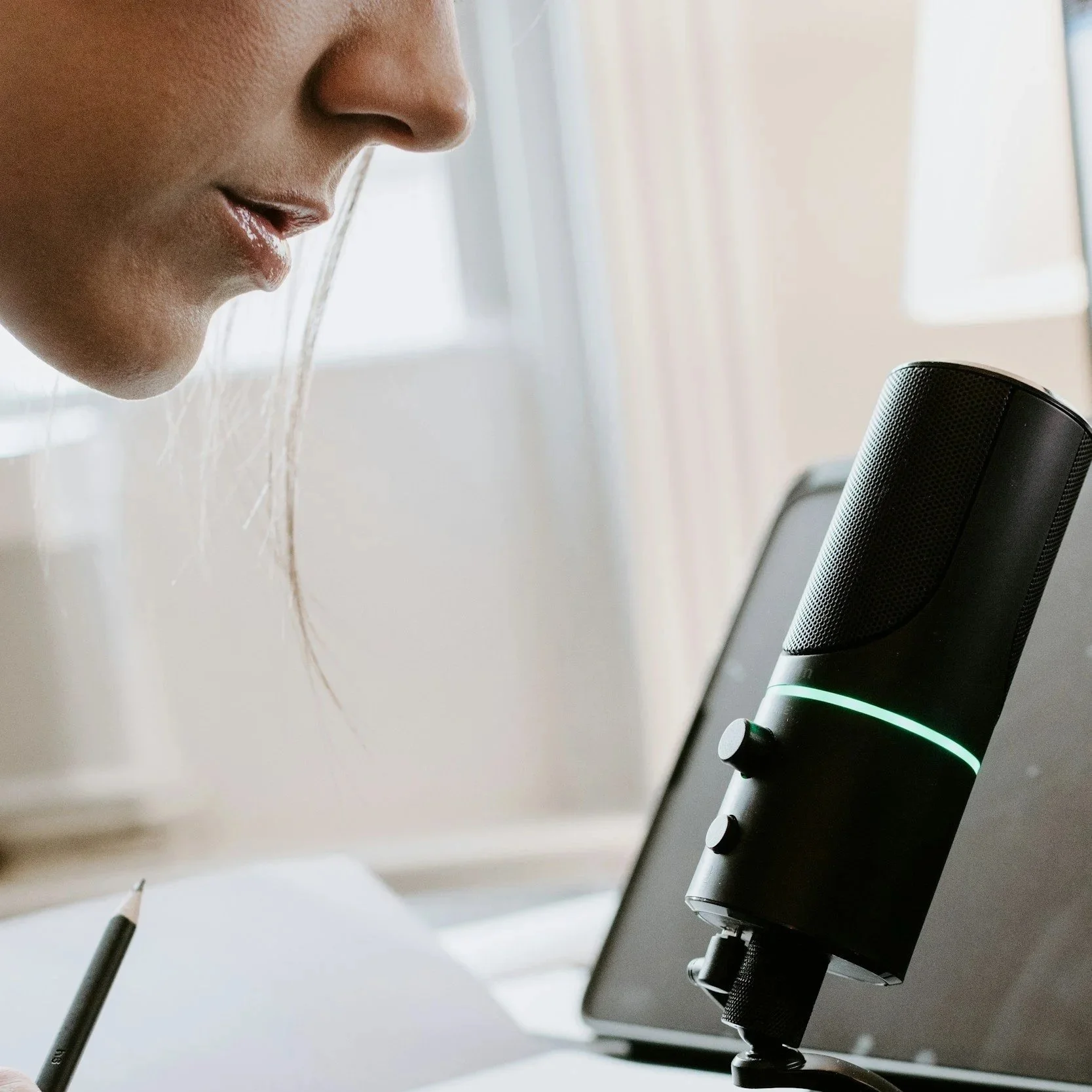 Close-up of a woman speaking into a black microphone with a green light, near a laptop on a desk