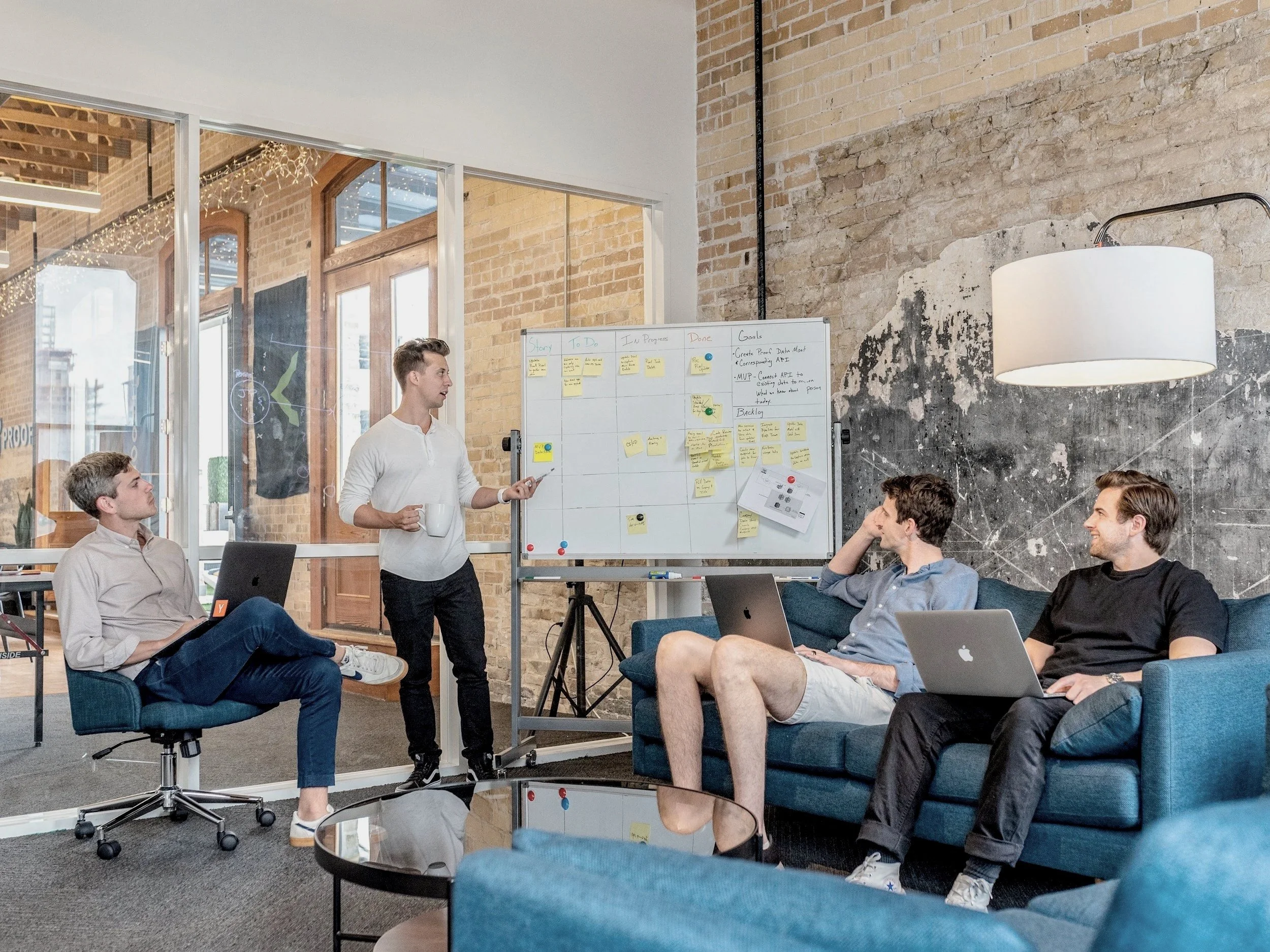Four young men in a modern office space participating in a presentation. One man is standing by a whiteboard with sticky notes, holding a mug, while three men seated with laptops listen attentively.