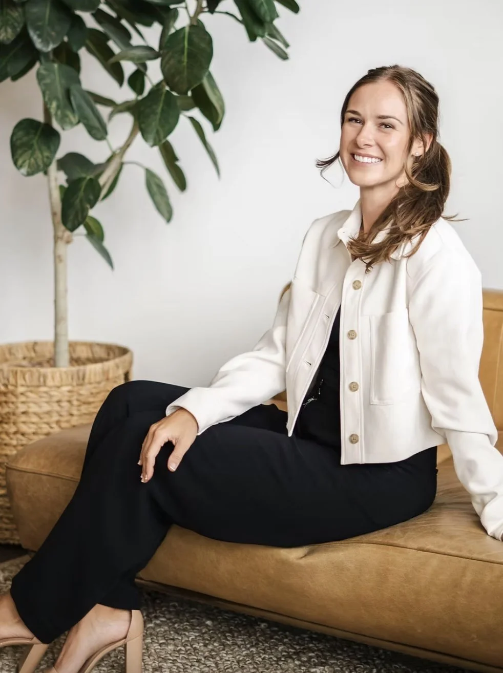 A woman sitting on a tan leather couch next to a large potted plant, smiling at the camera.