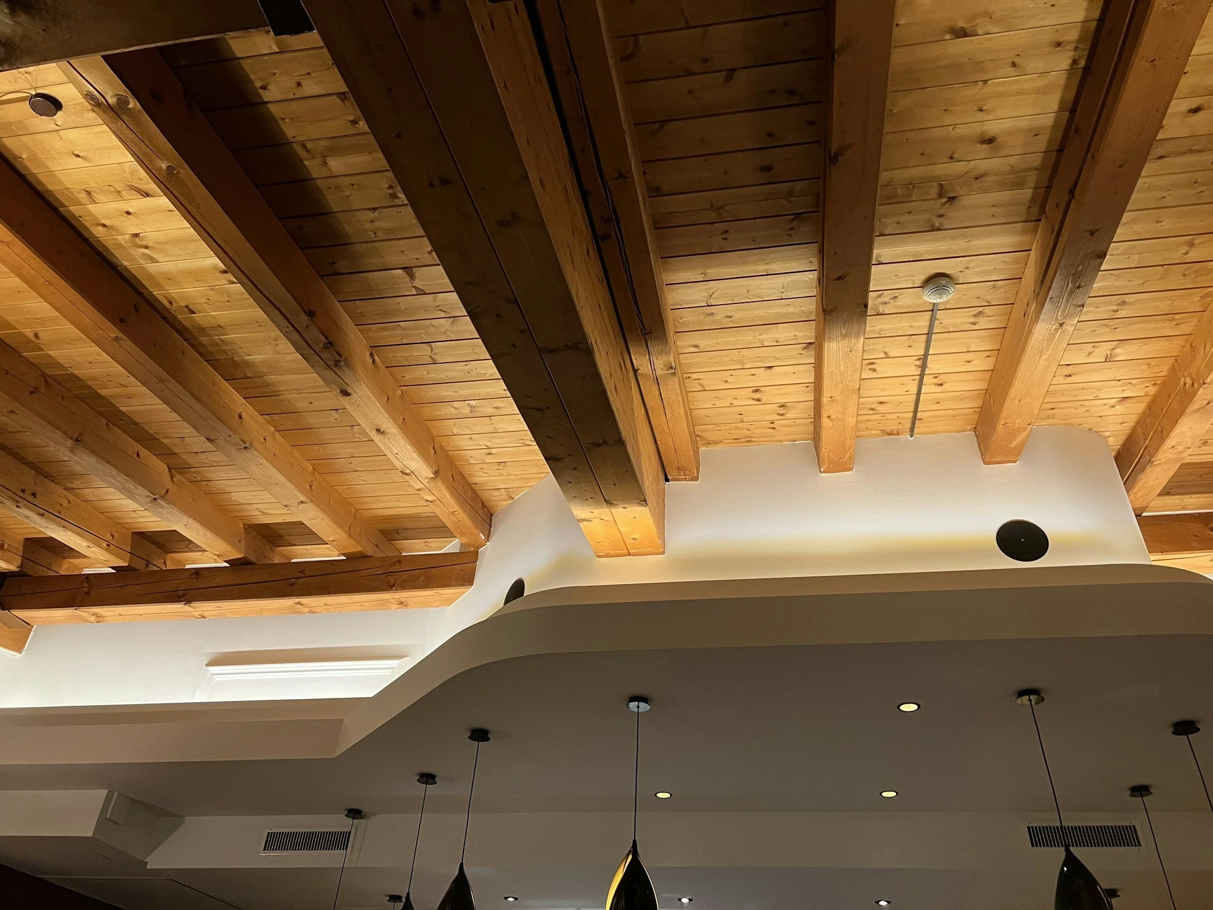 Wooden ceiling with exposed beams and a white wall with recessed lighting and hanging pendant lights.