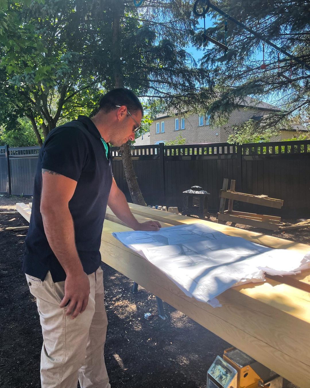 Man with sunglasses and a black shirt looking at architectural plans on a worktable outdoors, with trees and a black fence in the background.