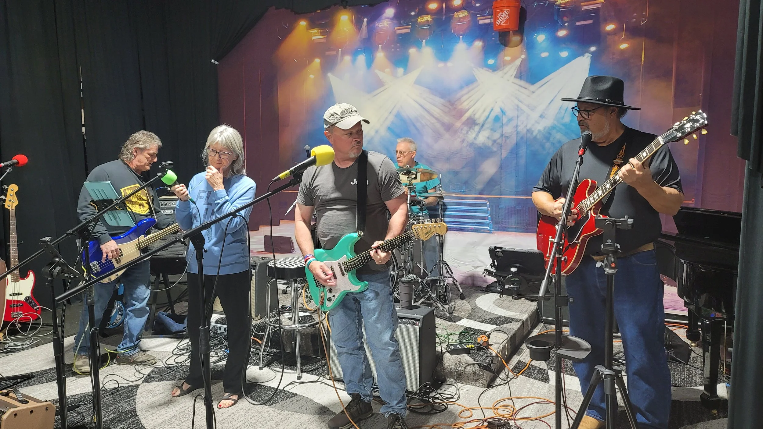A band of five musicians in a recording studio practicing or performing with guitars, drums, and microphones, with a colorful stage backdrop and stage lights.