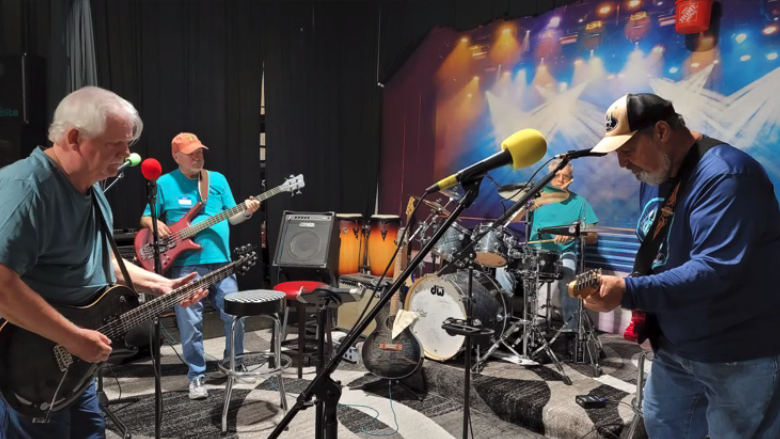 A group of musicians practicing or performing in a studio or rehearsal space with drums, guitars, and microphones.
