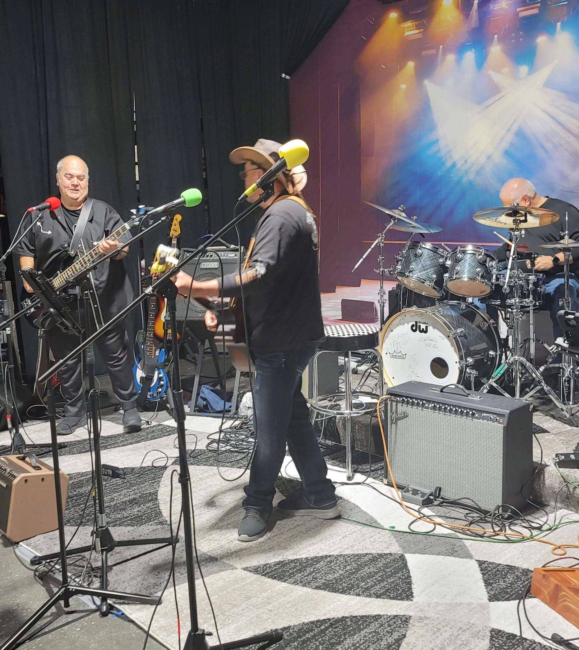 A band performing on stage with a drummer, bassist, and guitarist, with colorful stage lights and a dark backdrop.