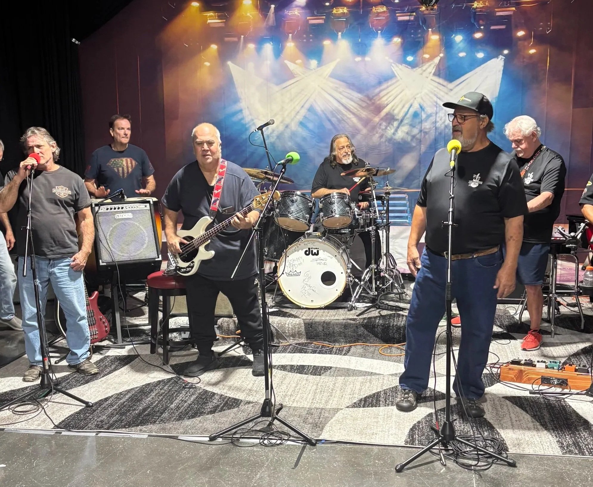 Group of musicians on stage with drums, guitars, and microphones, performing in a concert setting. Colorful stage lights shine down from above.