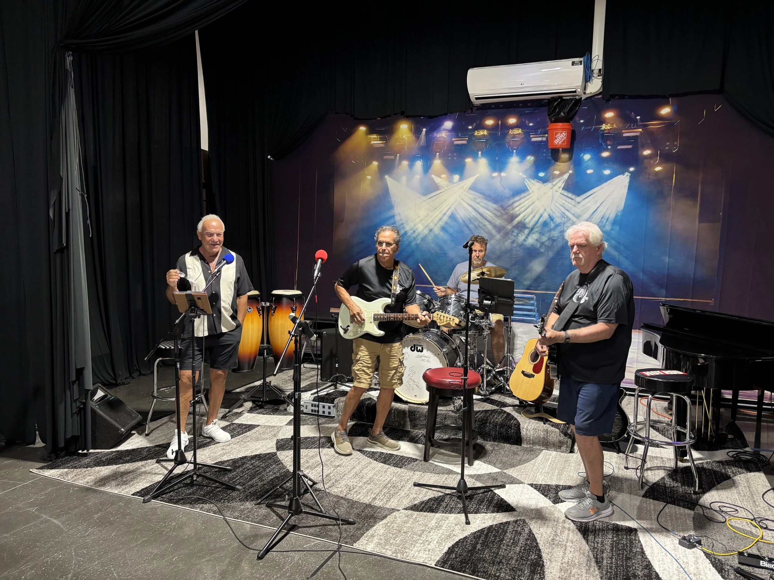 Four musicians performing on a stage with black curtains, various microphones, musical instruments including guitars, drums, congas, and a piano, under colorful stage lights and fog effects.