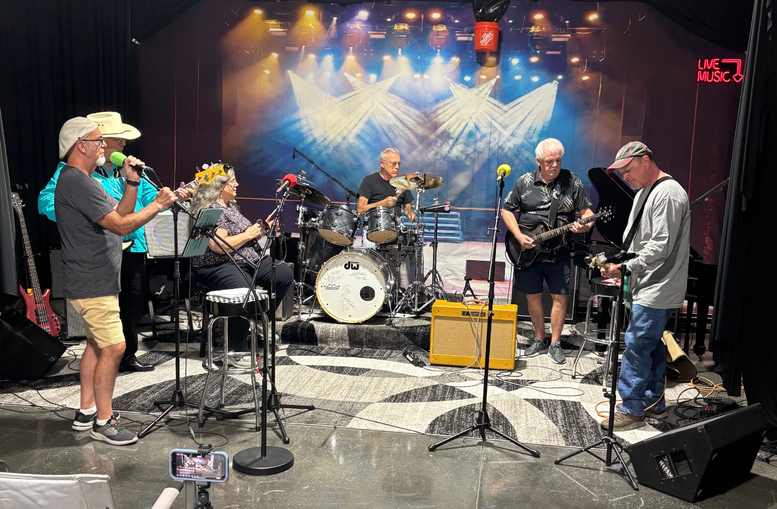 A band of six older adults performing on stage with various musical instruments, including guitars, drums, and a keyboard, under colorful stage lights with a 