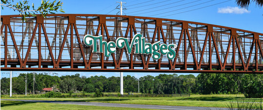A train bridge with a sign that reads 'The Villages' above a lush green grassy field and trees, under a blue sky.