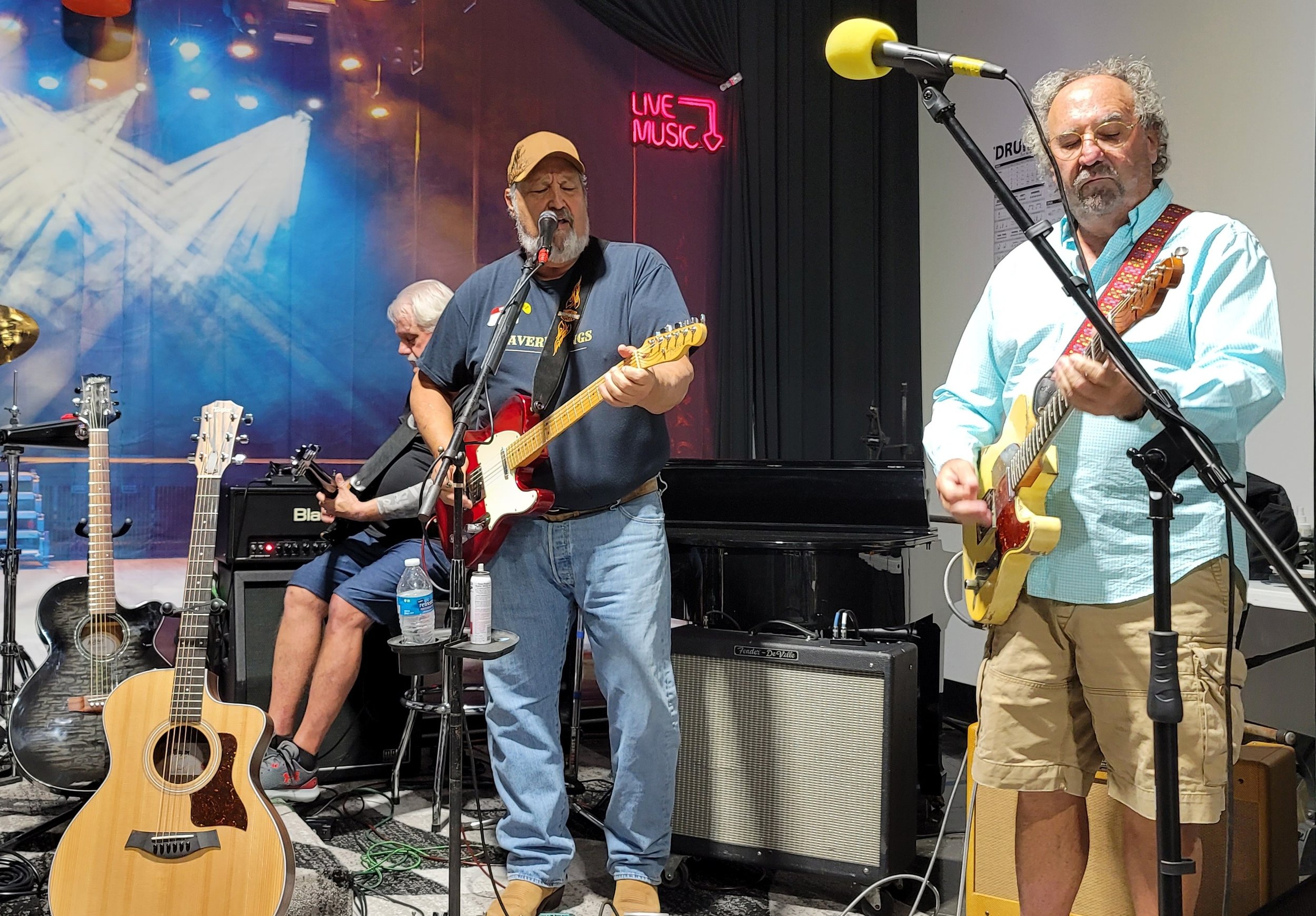 Three men playing guitars on a stage, with one singing into a microphone. The background features a blue stage curtain, a 'Live Music' neon sign, and stage lighting. Several additional guitars and amplifiers are visible.