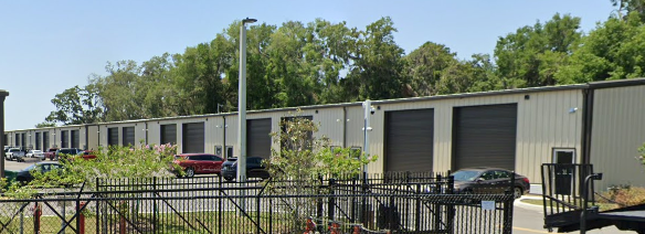 Row of units with black roll-up doors, parked cars, a chain-link fence, and surrounding trees under a blue sky. The external location of The Jam Zone