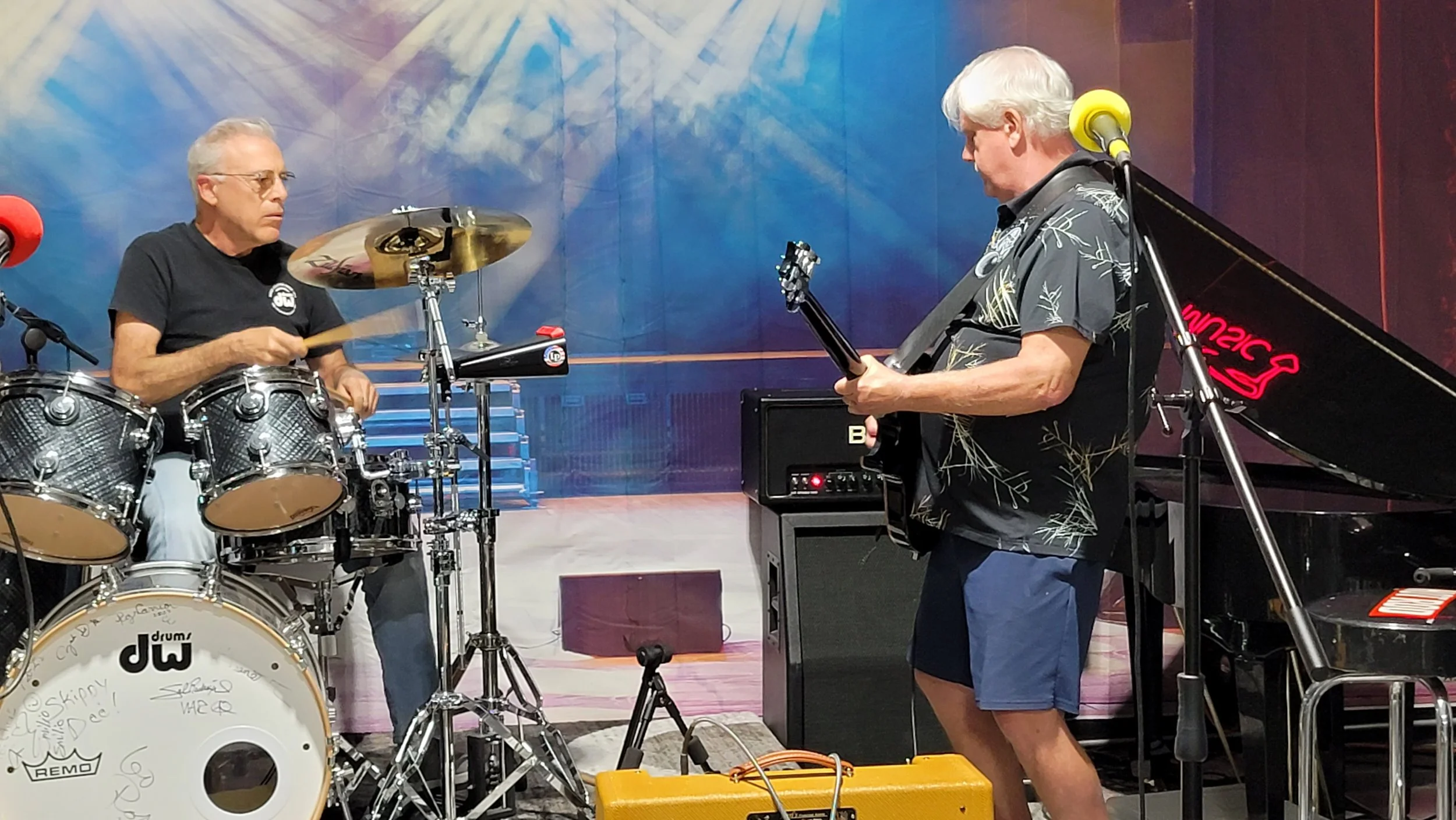 Two older men playing musical instruments in a music studio. One man is sitting at a drum set, hitting the drums with drumsticks. The other man is standing and playing an electric guitar. The background is a colorful wall and there is a piano on the 
