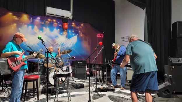 A group of older men rehearsing or performing on a small stage with musical instruments including a guitar, drums, and microphones with colorful stage lighting.