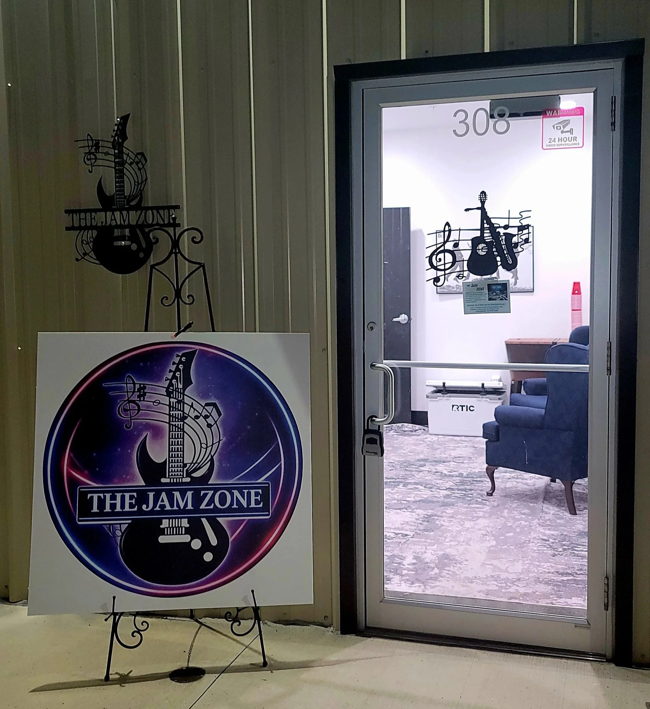 Entrance to a room called 'The Jam Zone,' with a sign on an easel and a glass door showing a music-themed wall decoration of a guitar and musical notes inside.