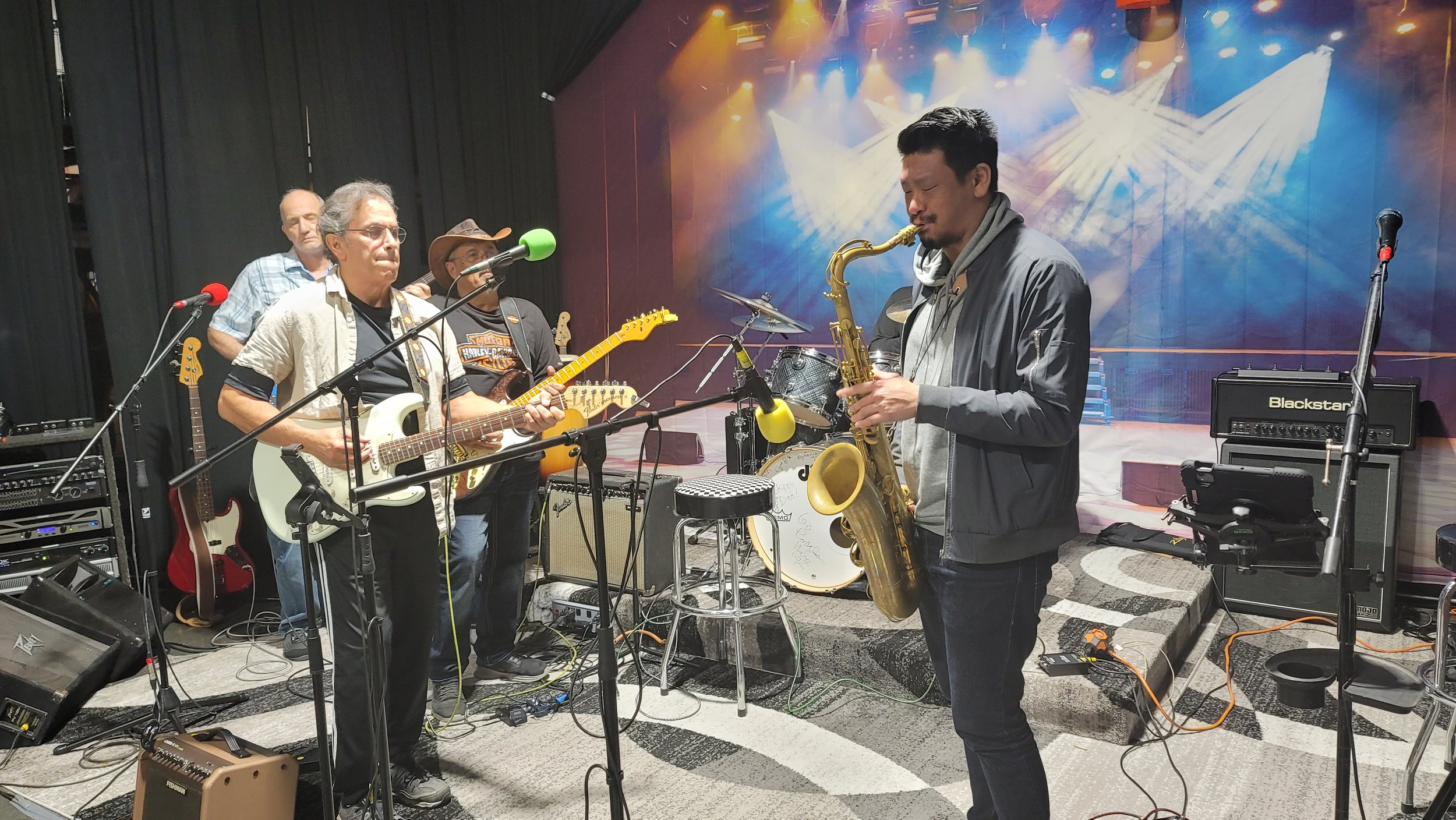 A band practicing on stage with four musicians, including a man playing an electric guitar, another playing a saxophone, a percussionist, and another musician in the background, with microphones, amplifiers, and musical equipment around.