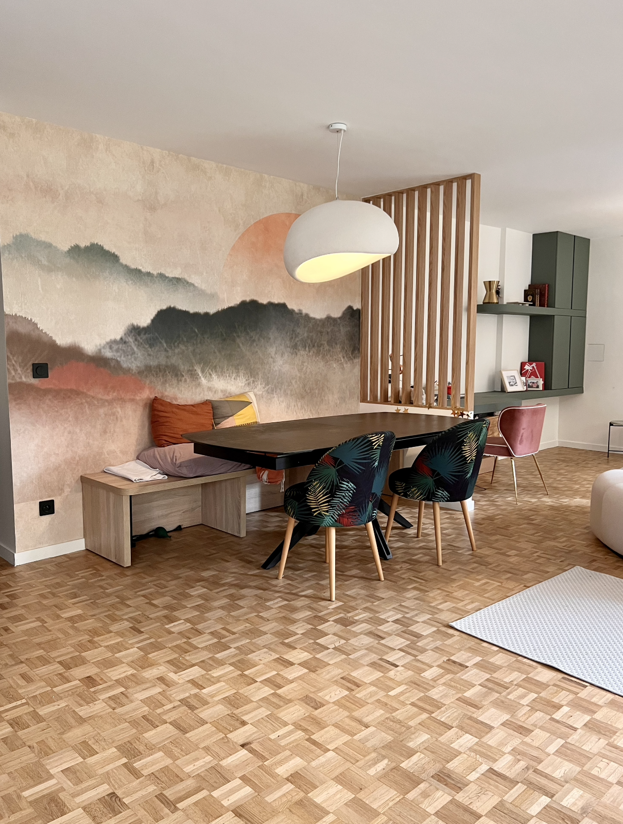 Living room with a wooden parquet floor, a mural of mountains and a sun on the wall, a black dining table with floral-patterned chairs, a padded chair, and a hanging white lamp.