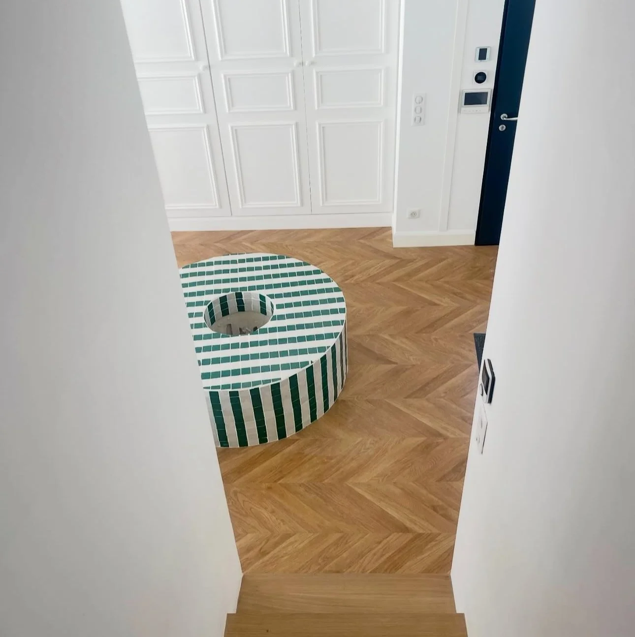 View of a modern interior with wooden herringbone flooring, white paneled walls, a white door, and a colorful mosaic round seating area with green, white, and black tiles.