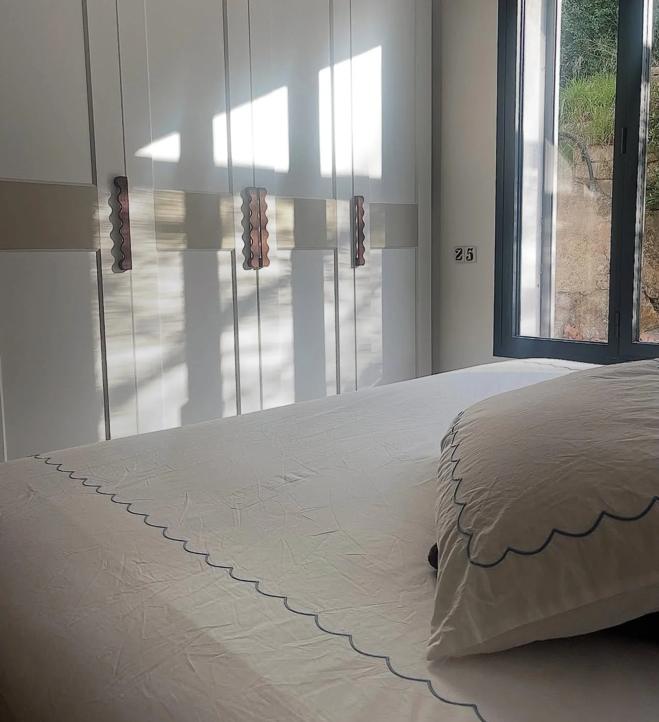 A bedroom with a bed in the foreground, white bedding with decorative stitching, a white wardrobe with vertical panels and handles, a black-framed window slightly open, and sunlight casting shadows on the wardrobe.