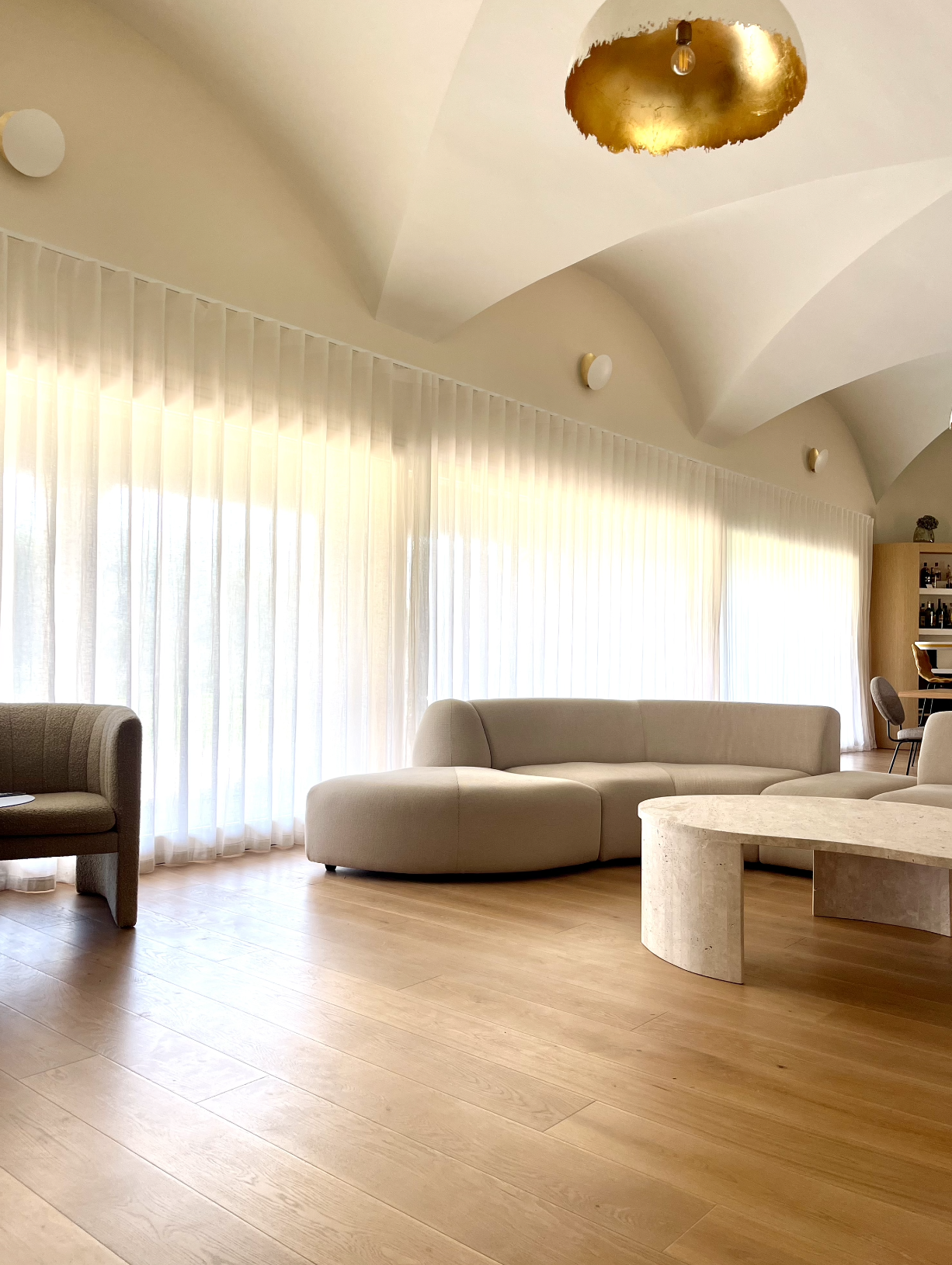 A modern living room with light-colored curved sofas, sheer curtains, wooden flooring, and a gold ceiling light fixture.