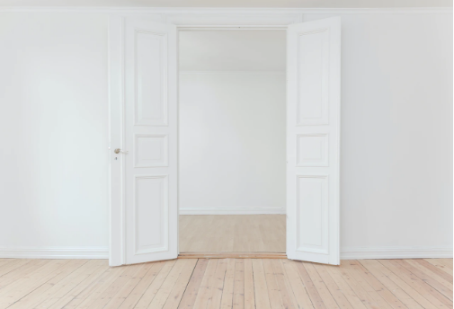 Empty white room with double doors opening to another room with similar white walls and a light wood floor.