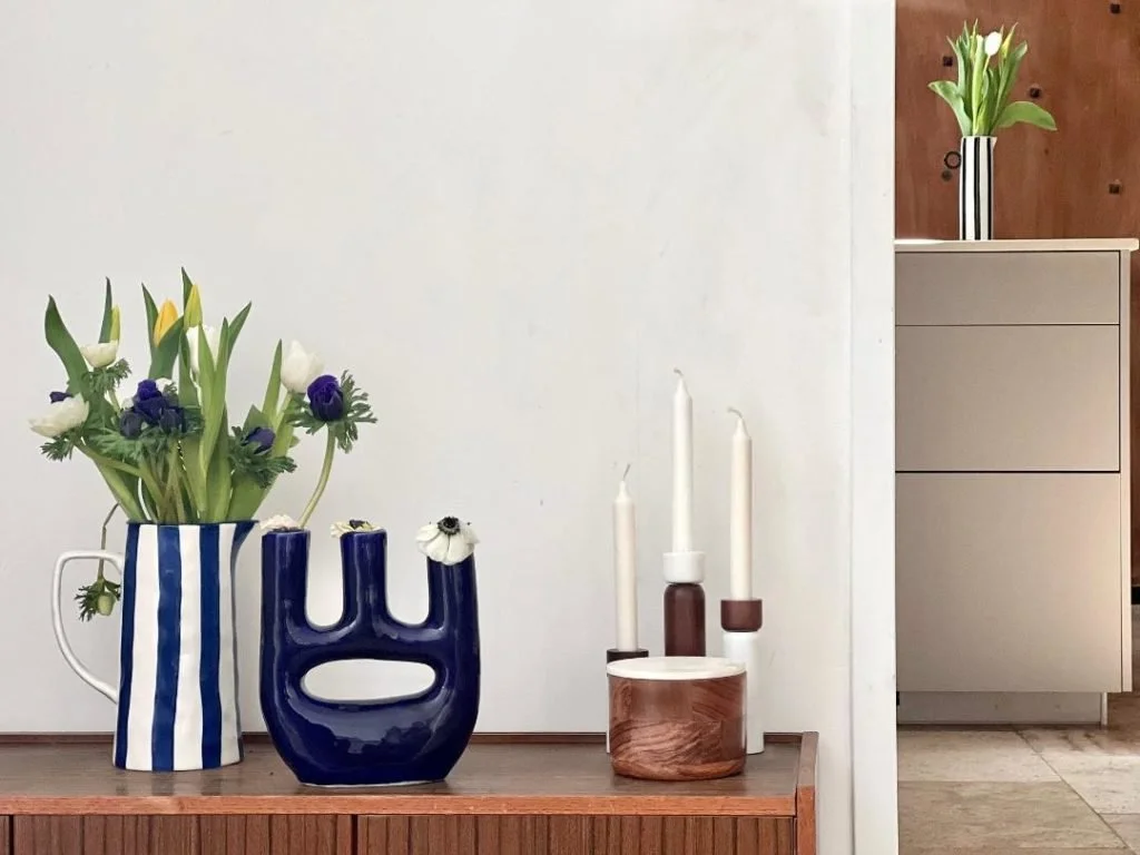 Vases with flowers and candles on wooden furniture in a room with minimal decor.