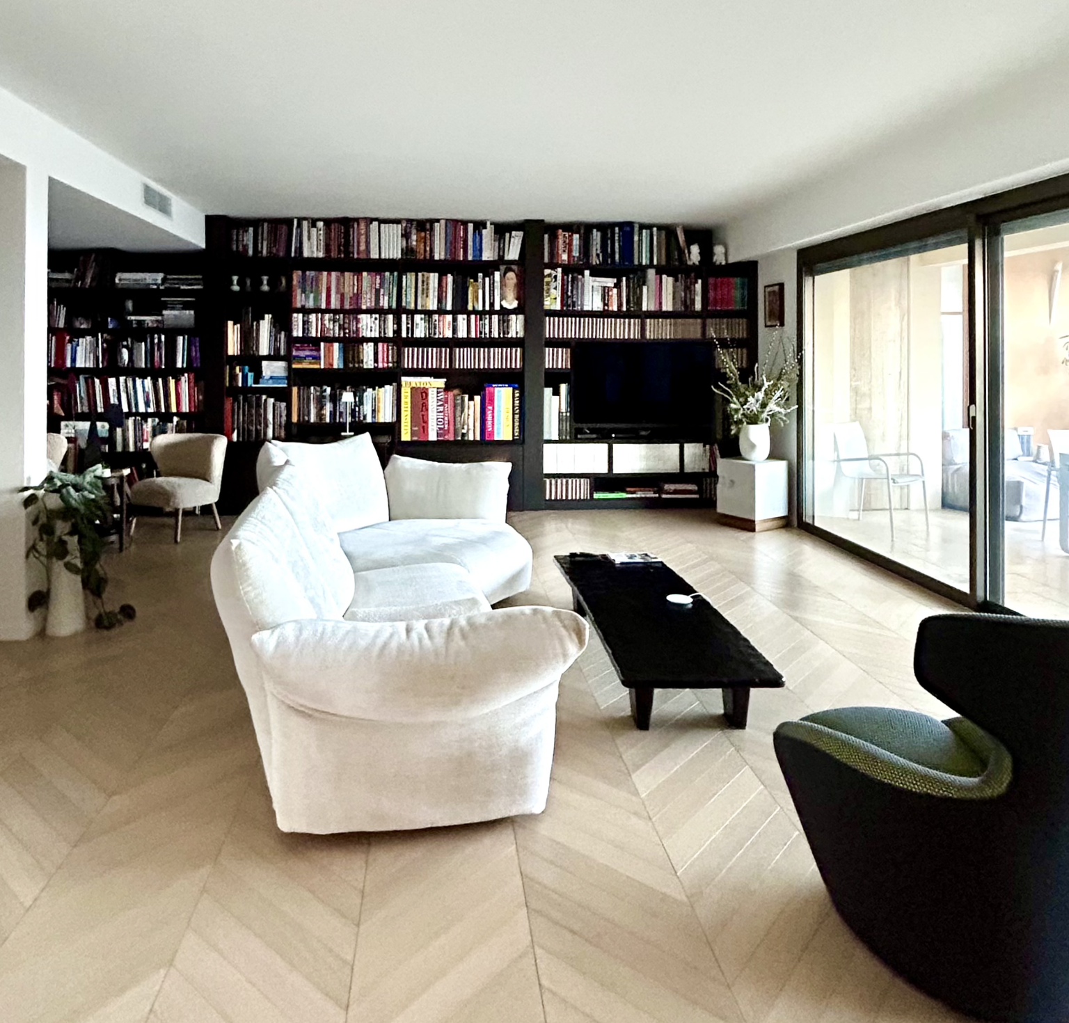 Living room with white sofa, black armchair, wooden coffee table, and large bookshelf filled with books, with sliding glass doors leading to a balcony with chairs.
