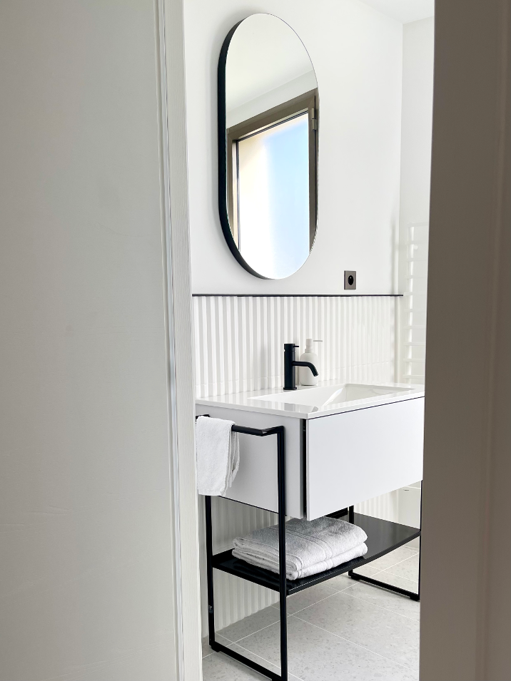 Modern bathroom vanity with a black faucet, white countertop, oval mirror, and a towel rack with towels, in a minimalist style.